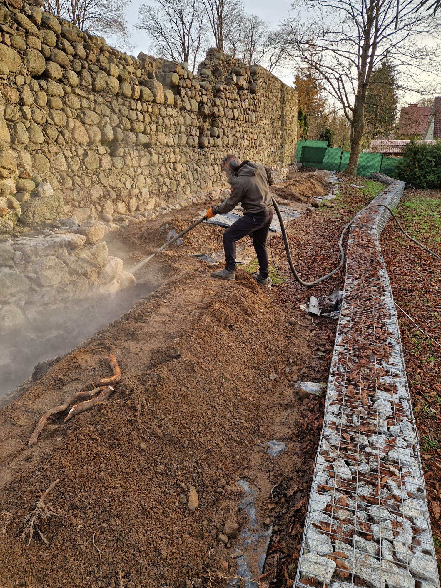 Czyszczenie kamiennej ściany strumieniem pod ciśnieniem przez pracownika w ogrodzie. Gabiony w tle.
