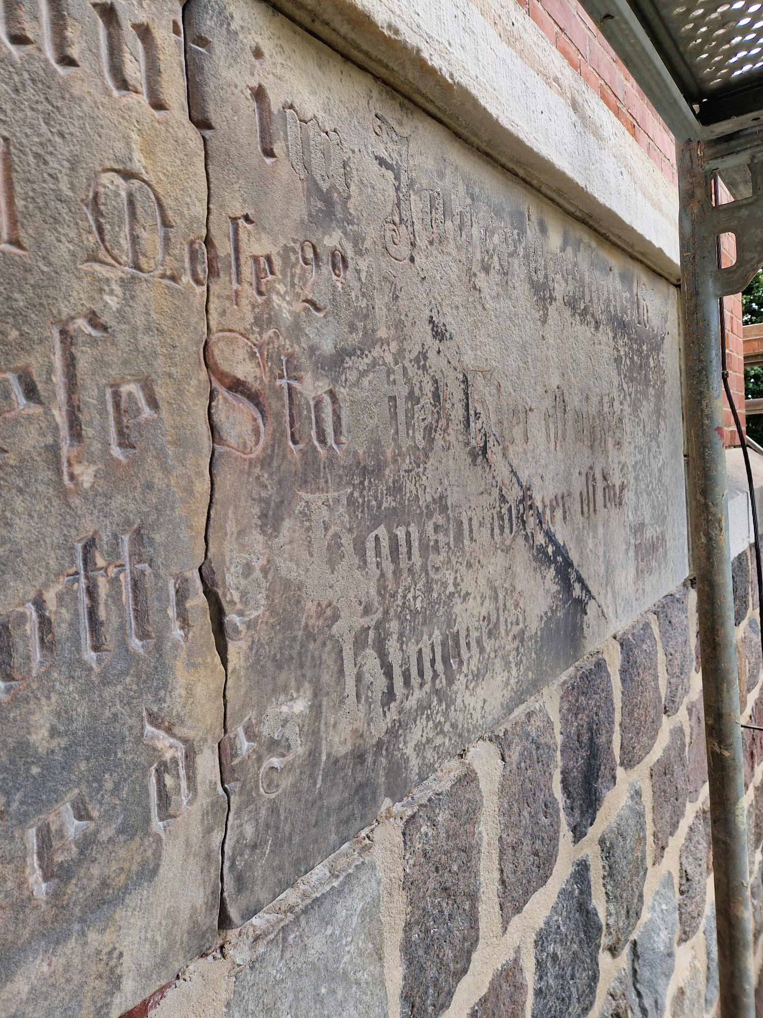 Kamienna ściana z wyrytymi inskrypcjami, częściowo odnowiona, z widocznym rusztowaniem. Detal elewacji zabytkowego budynku w trakcie prac konserwatorskich.