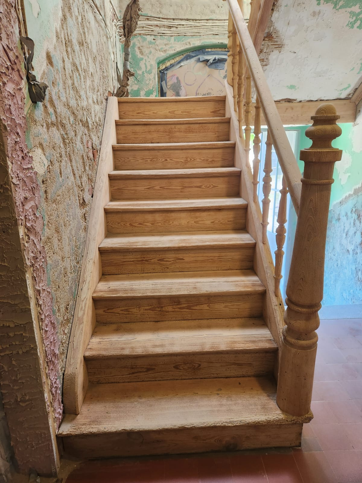 Drewniane schody z balustradą w odrestaurowywanym wnętrzu. Widoczne ślady starej farby na ścianach i elementy dekoracyjne.