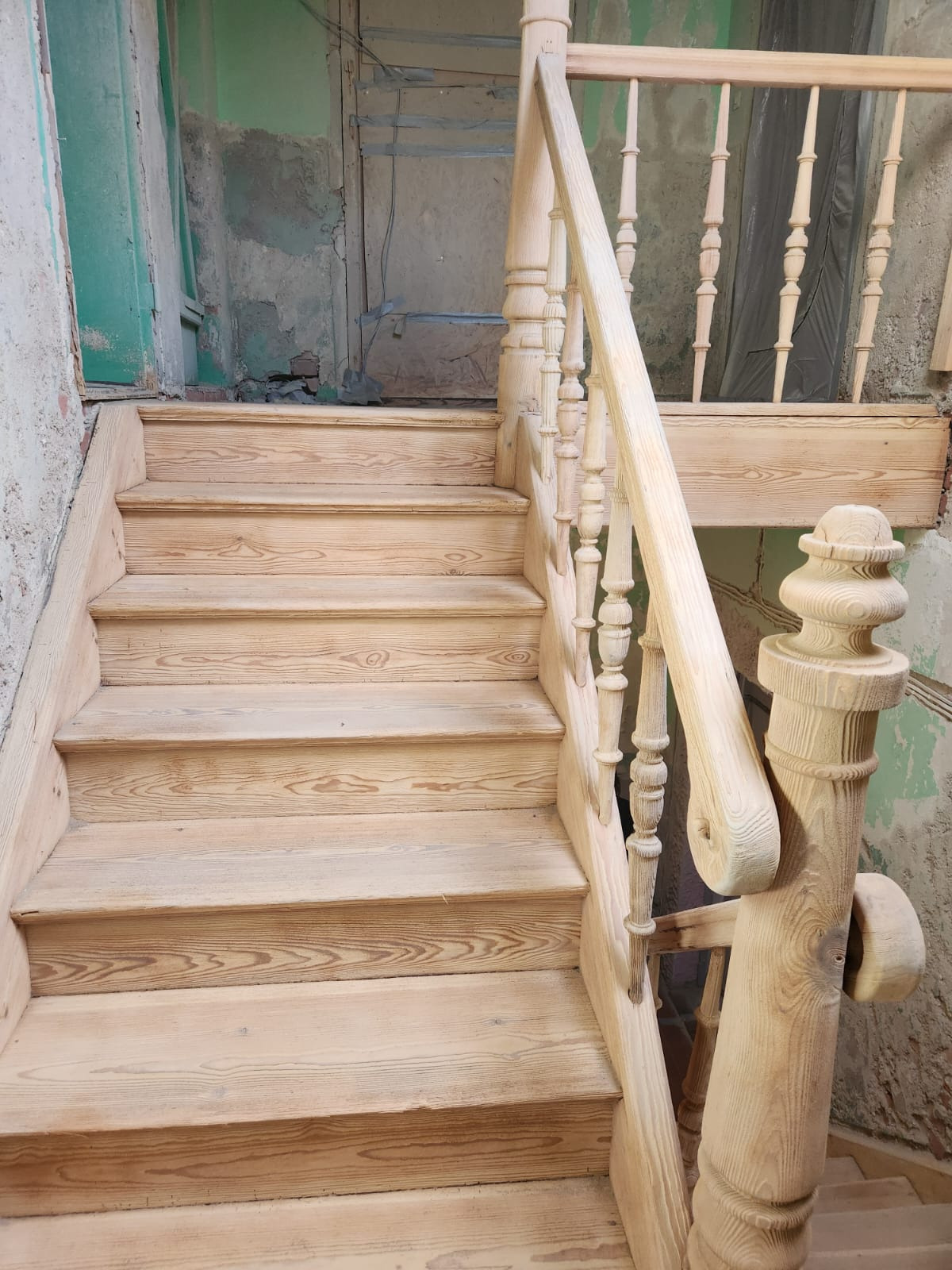 Drewniane schody z balustradą w surowym stanie, widoczne słoje drewna, fragment ściany z zieloną farbą w tle. Schody w trakcie renowacji lub montażu.