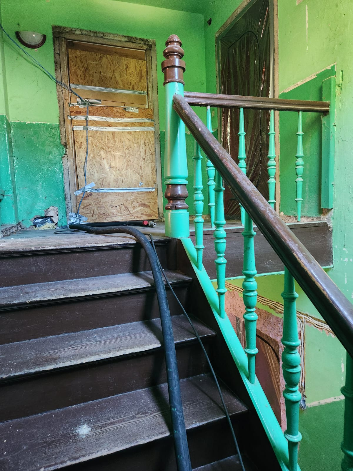 Klatka schodowa w budynku z lat 70. z zielonymi ścianami i drewnianymi schodami przed remontem. Widoczne zabezpieczone drzwi i rura od odkurzacza.