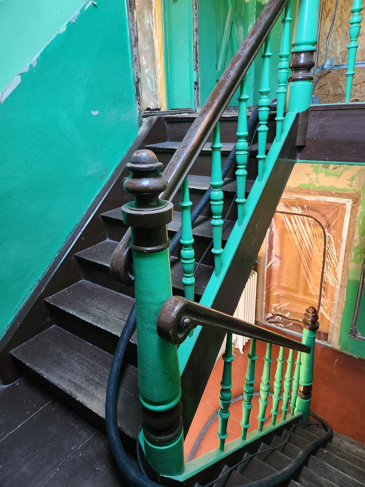 Drewniane schody z zieloną balustradą w starym domu, widok z góry. Elementy schodów częściowo zakryte folią ochronną podczas prac remontowych. Widoczne ślady użytkowania.