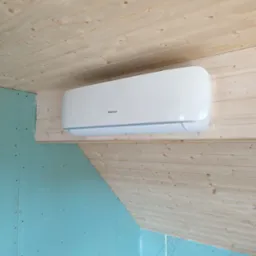 Biały klimatyzator Hisense zamontowany na drewnianej ścianie pod skośnym drewnianym sufitem, ściana boczna z płyty gipsowej w kolorze turkusowym.
