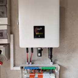 Biały inwerter fotowoltaiczny marki FOX ESS zamontowany na szarej ścianie, z widoczną skrzynką bezpieczników z pomarańczowymi i zielonymi elementami oraz przewodami. Na wyświetlaczu inwertera...