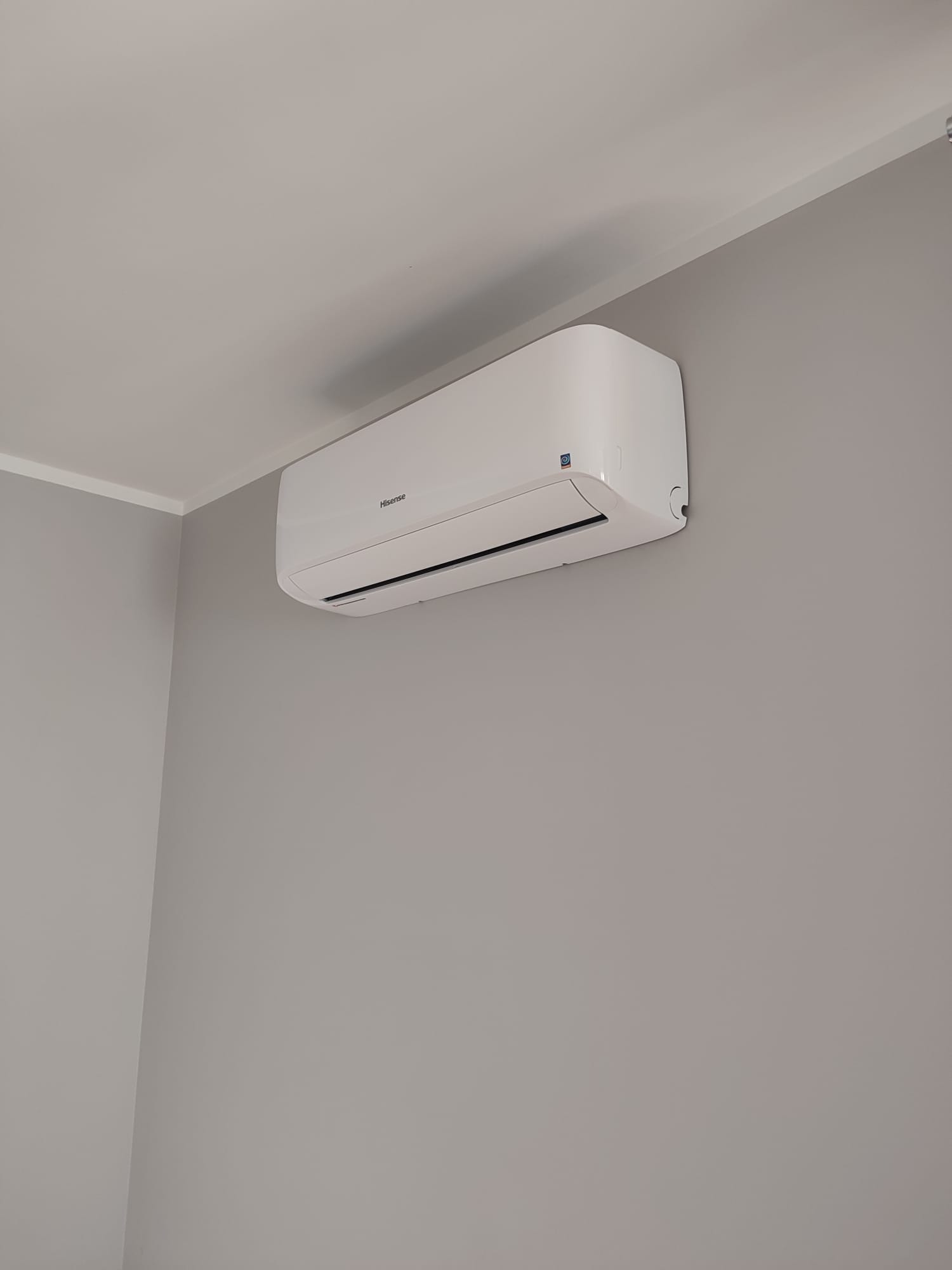 Biały klimatyzator marki Hisense zamontowany na szarej ścianie w rogu pomieszczenia, widoczne elementy instalacji elektrycznej.