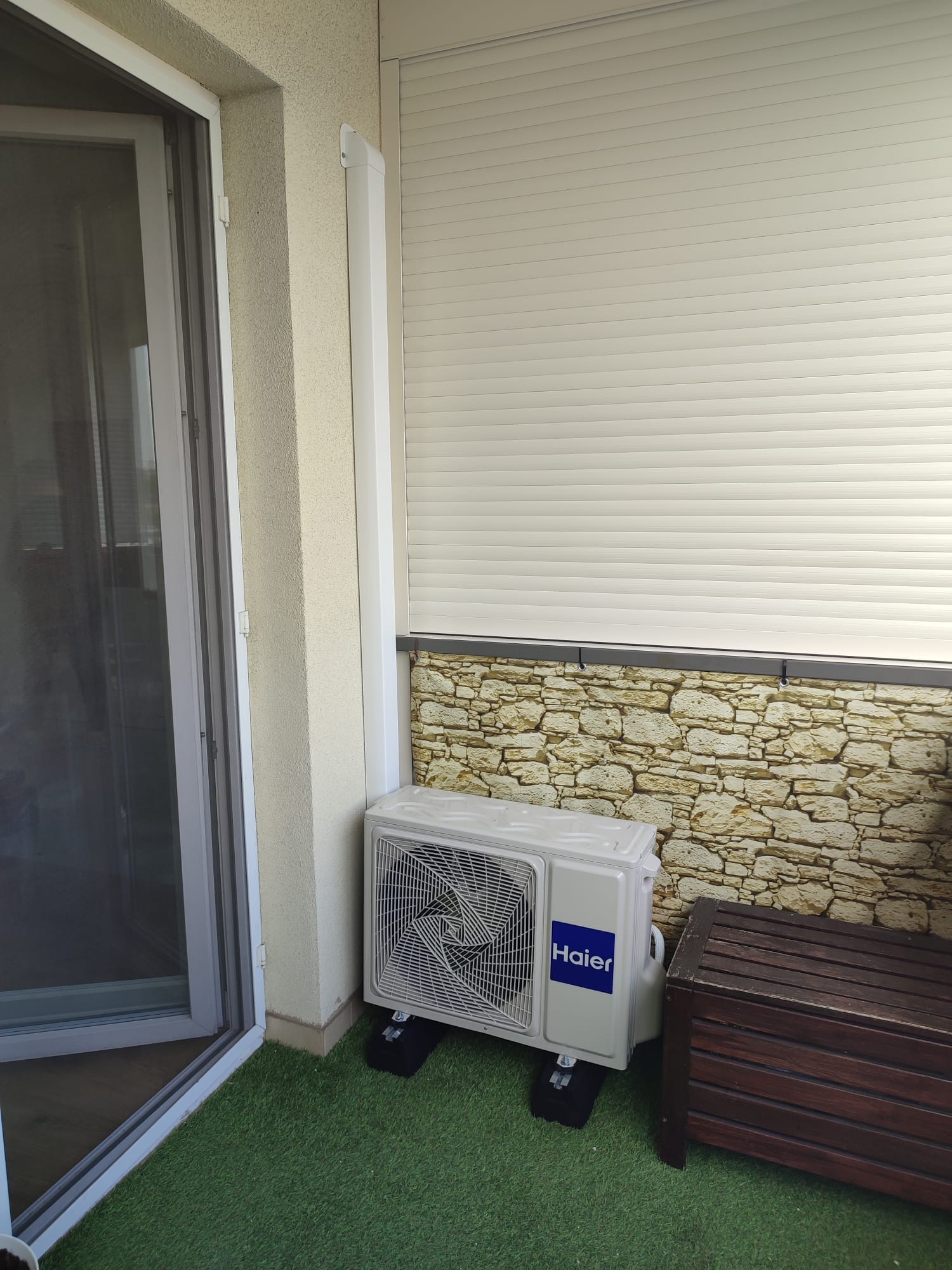 Zewnętrzna jednostka klimatyzatora Haier na balkonie z imitacją trawy, obok drewniana skrzynia i ściana wyłożona kamieniem dekoracyjnym pod żaluzjami.