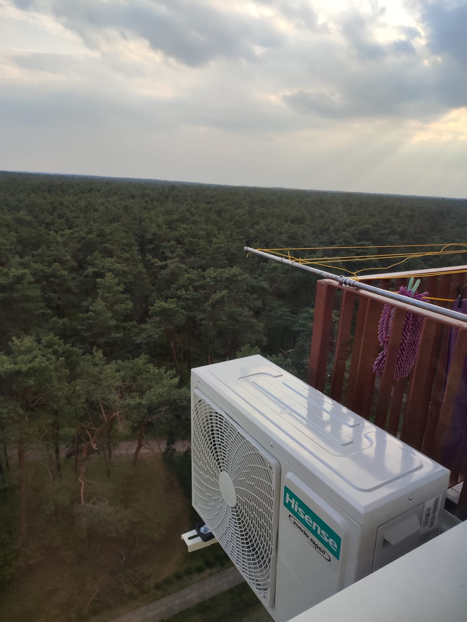 Biała jednostka zewnętrzna klimatyzatora Hisense zamontowana na balkonie z widokiem na rozległy las i pochmurne niebo z promieniami słońca.