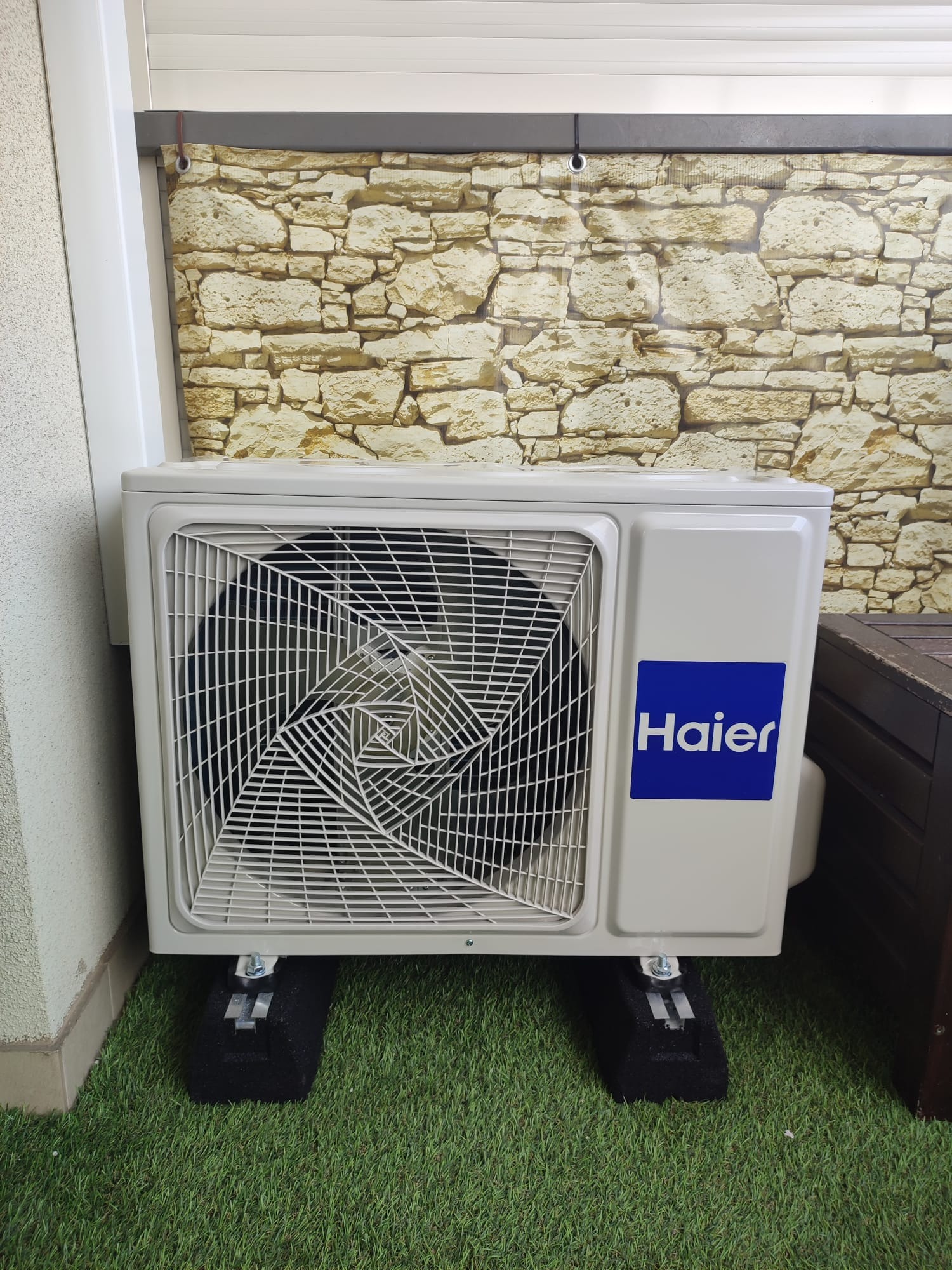 Zewnętrzna jednostka klimatyzacji Haier na zielonej sztucznej trawie, zamontowana na czarnych podkładkach antywibracyjnych, z tłem imitującym ścianę z kamienia.
