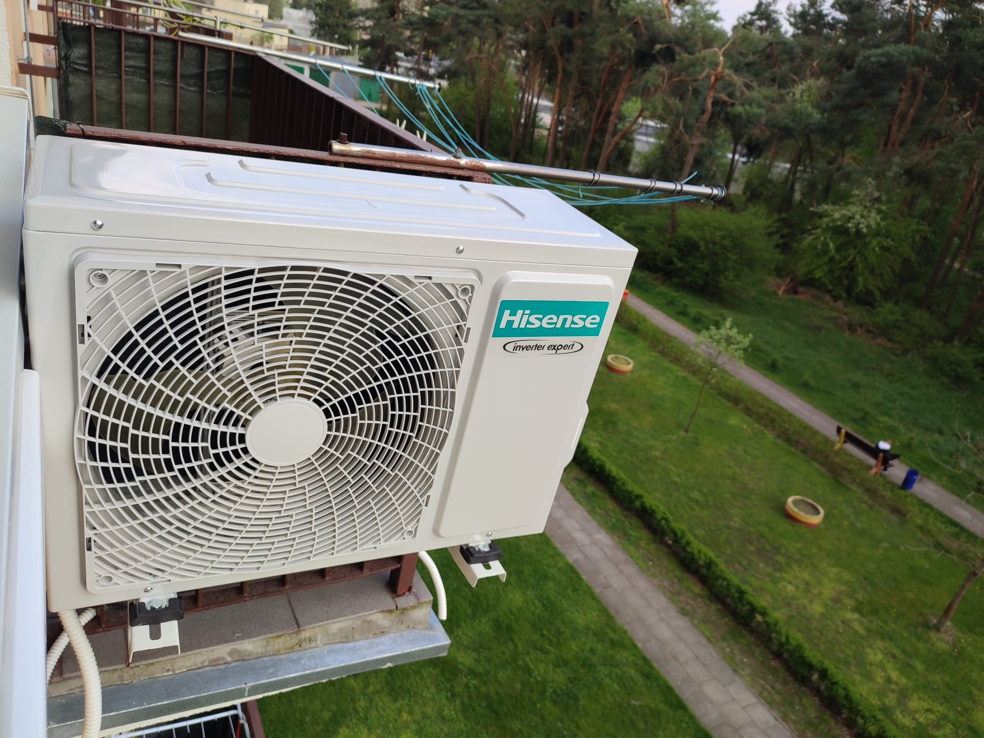 Biała jednostka zewnętrzna klimatyzacji marki Hisense zamontowana na balkonie, widok z góry na trawnik z chodnikiem i drzewami w tle.
