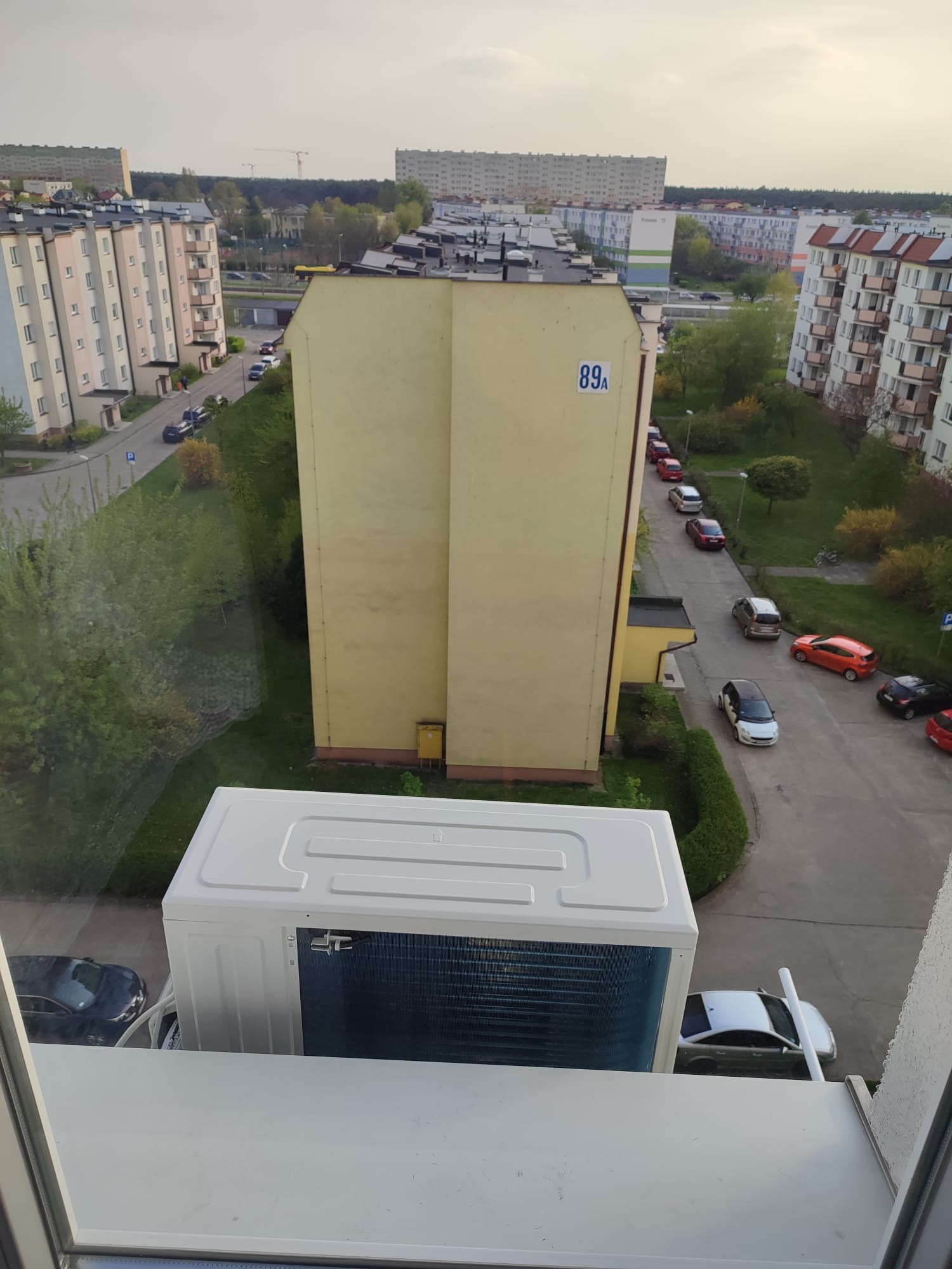 Zewnętrzna jednostka klimatyzacji zamontowana na balkonie z widokiem na osiedle bloków mieszkalnych, widoczne uliczki i zieleń.