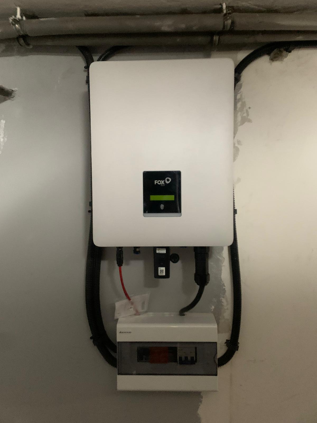 Biały inwerter z logo FOX podłączony do skrzynki elektrycznej na szarej ścianie, widoczne przewody i rurki instalacyjne.