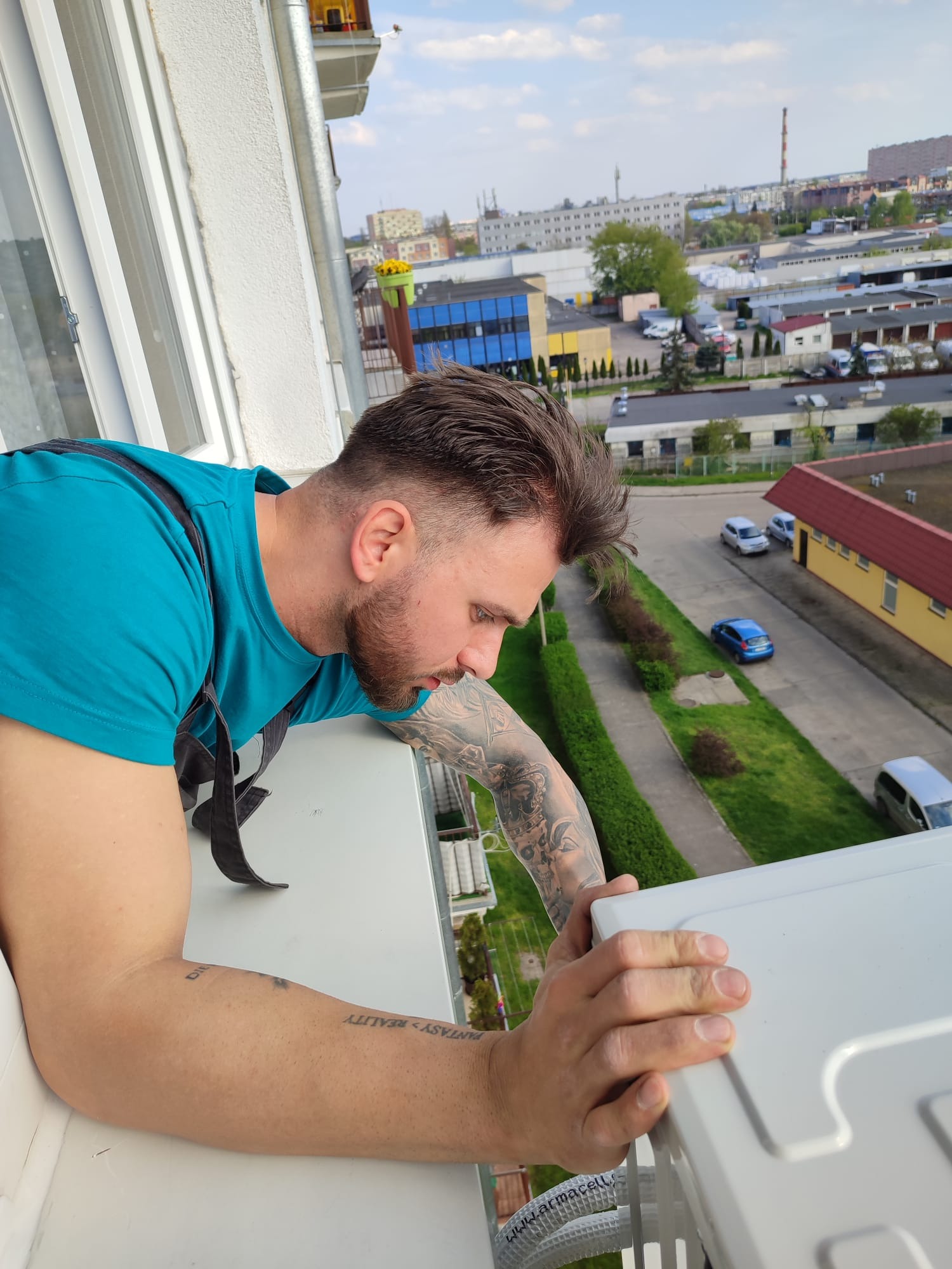 Mężczyzna w niebieskiej koszulce, z tatuażami na ramieniu, instaluje zewnętrzną jednostkę klimatyzacji na balkonie, z widokiem na miejski krajobraz w tle.