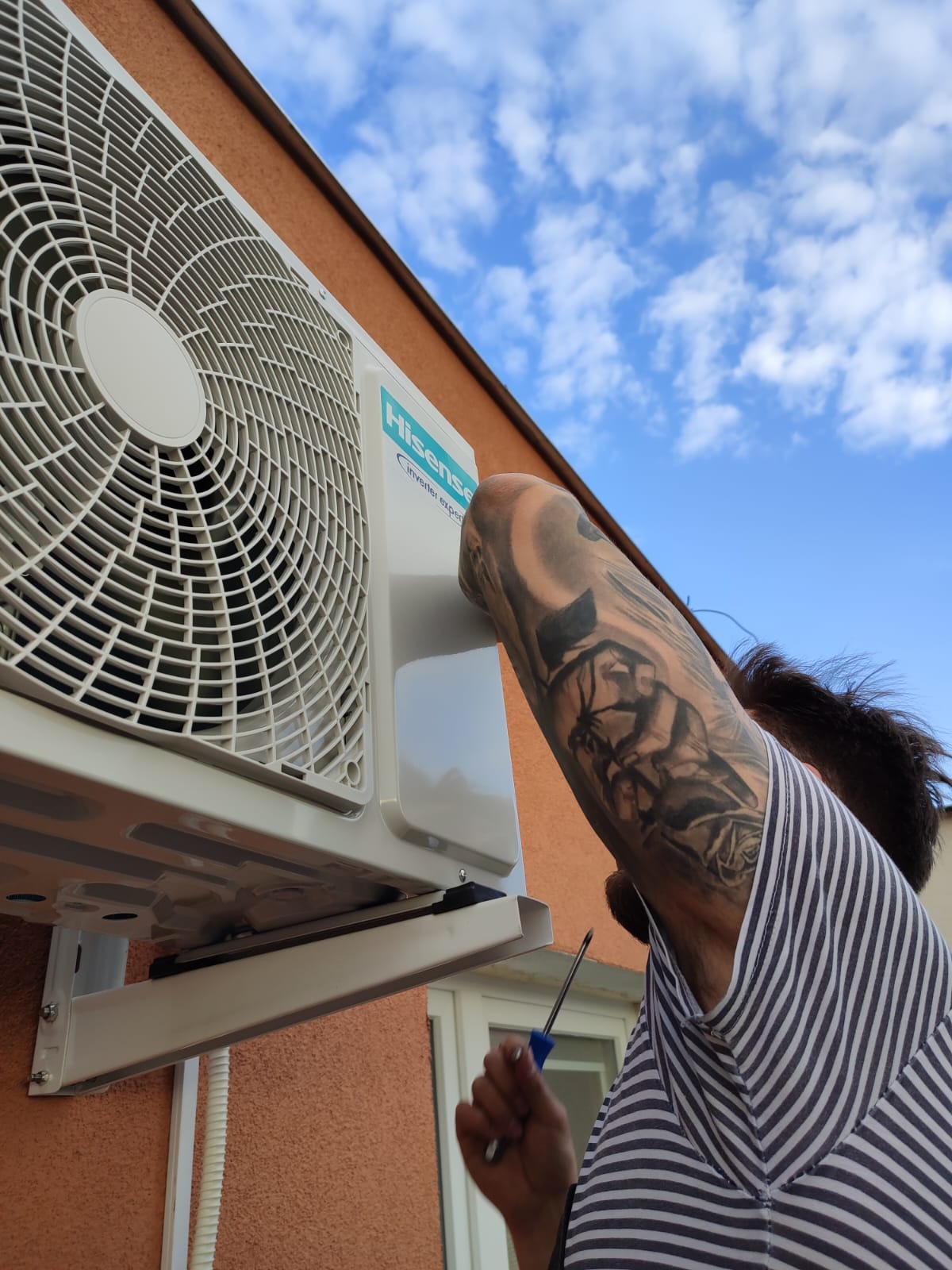 Instalacja zewnętrznej jednostki klimatyzacji Hisense na pomarańczowej ścianie budynku, widok z dołu, ramię instalatora z tatuażem, niebieskie niebo w tle.