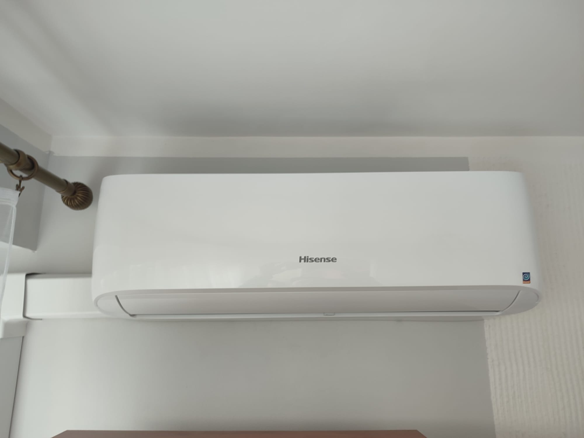 Biała jednostka wewnętrzna klimatyzacji marki Hisense zamontowana na ścianie, widoczne rury instalacyjne w białej osłonie i fragment karnisza.