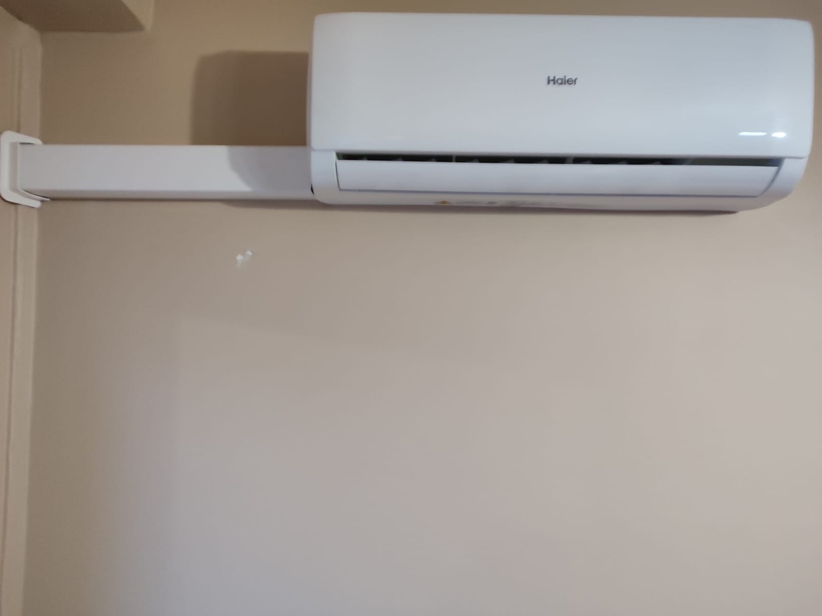 Biała jednostka klimatyzacyjna Haier zamontowana na kremowej ścianie, z widocznym mocowaniem na ścianie po lewej stronie.