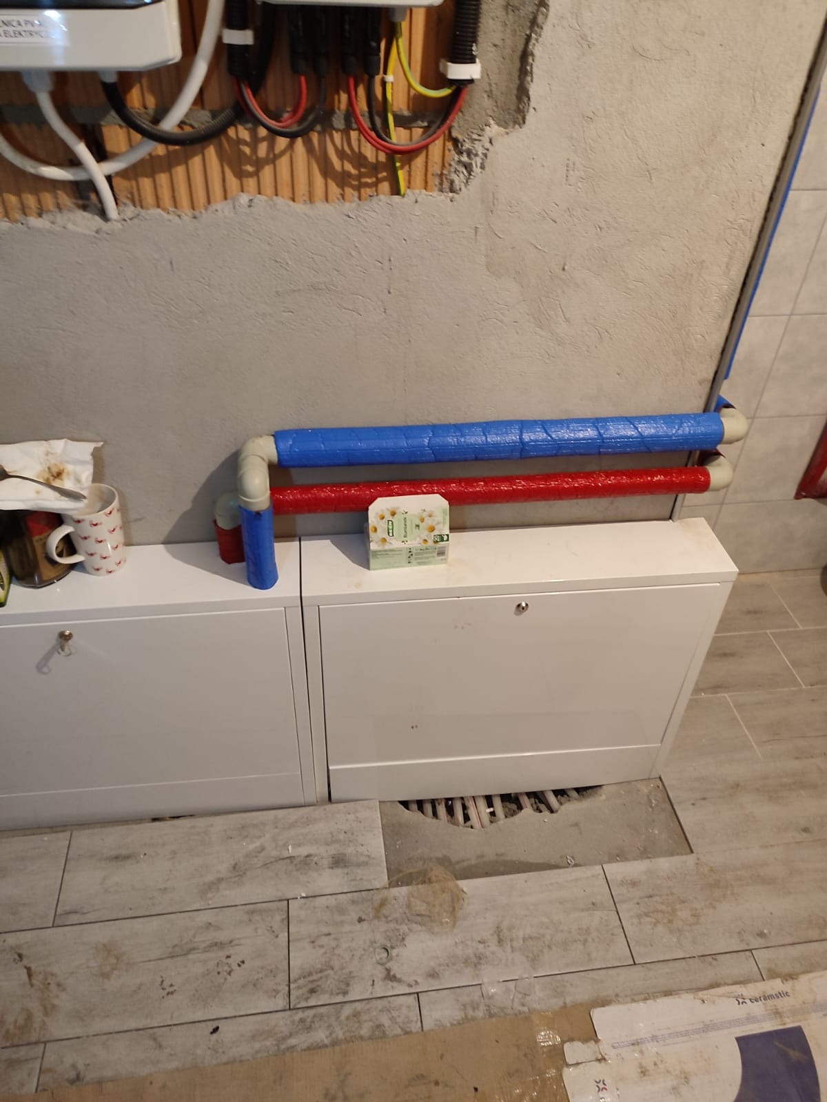 Fragment instalacji z rurami w izolacji w kolorach niebieskim i czerwonym, prowadzącymi nad białymi szafkami w surowym wnętrzu z niedokończonymi płytkami podłogowymi i widocznymi przewodami...