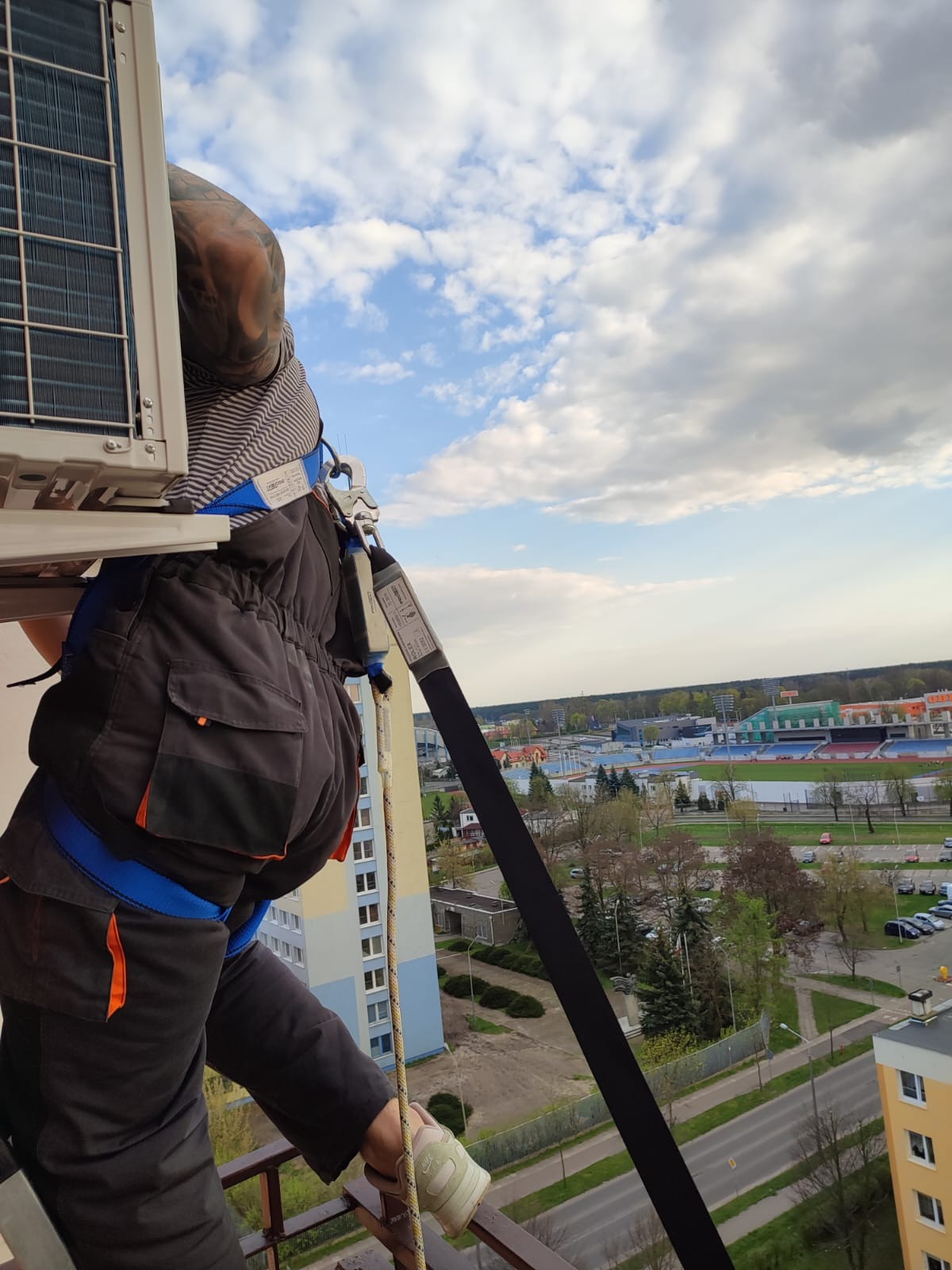 Instalator w uprzęży montuje zewnętrzną jednostkę klimatyzacji na balkonie wysokiego budynku, widok na miasto w tle.