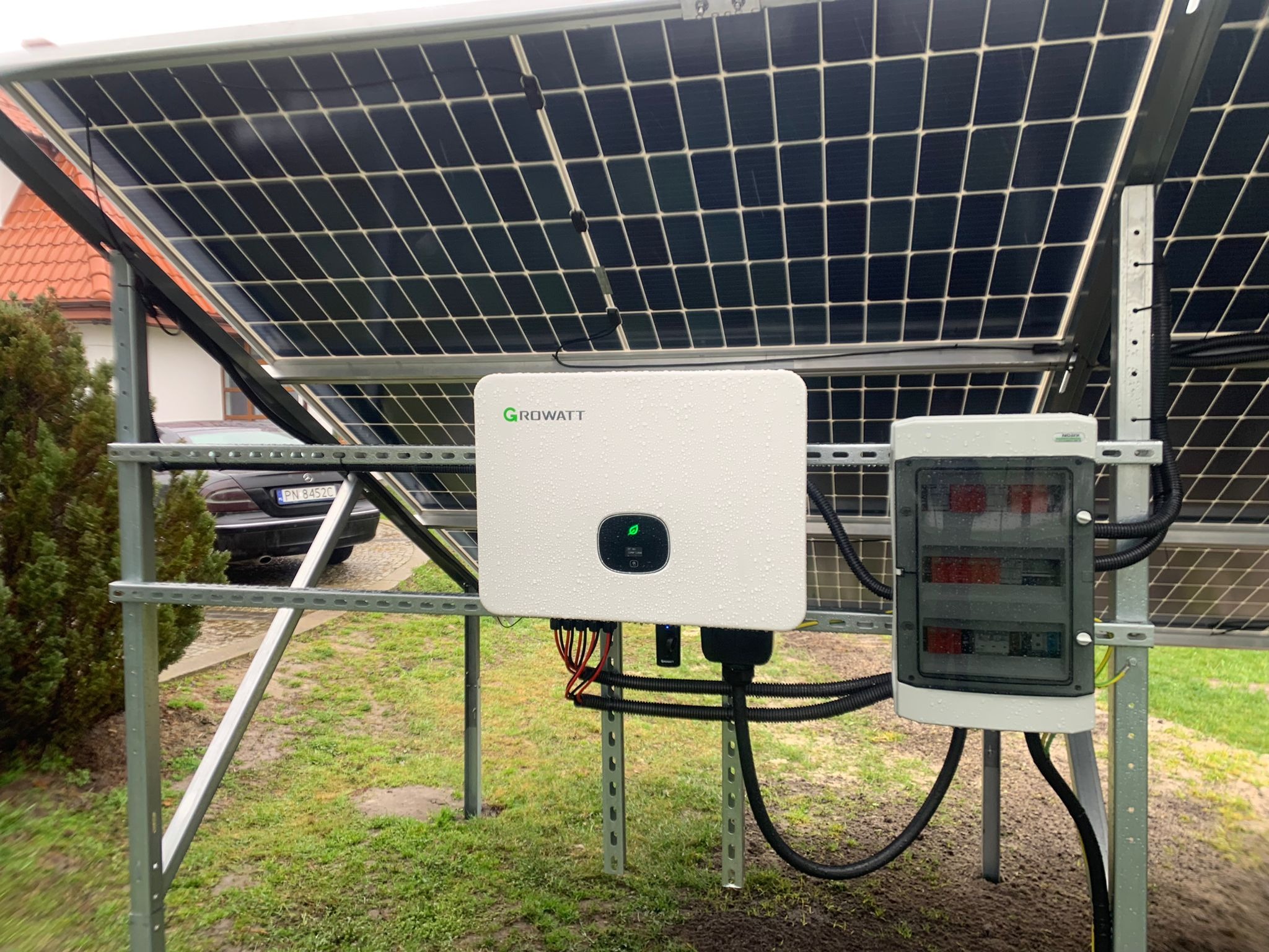 Panel fotowoltaiczny na metalowej konstrukcji, biały inwerter Growatt z kroplami deszczu, skrzynka elektryczna, trawa i fragment samochodu w tle.