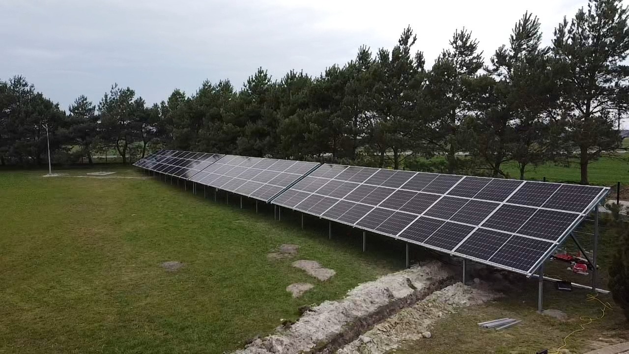 Rozległa instalacja paneli słonecznych na trawiastym terenie z rzędem drzew w tle, widoczny wykop pod przyszłe instalacje, elementy montażowe i przewody.
