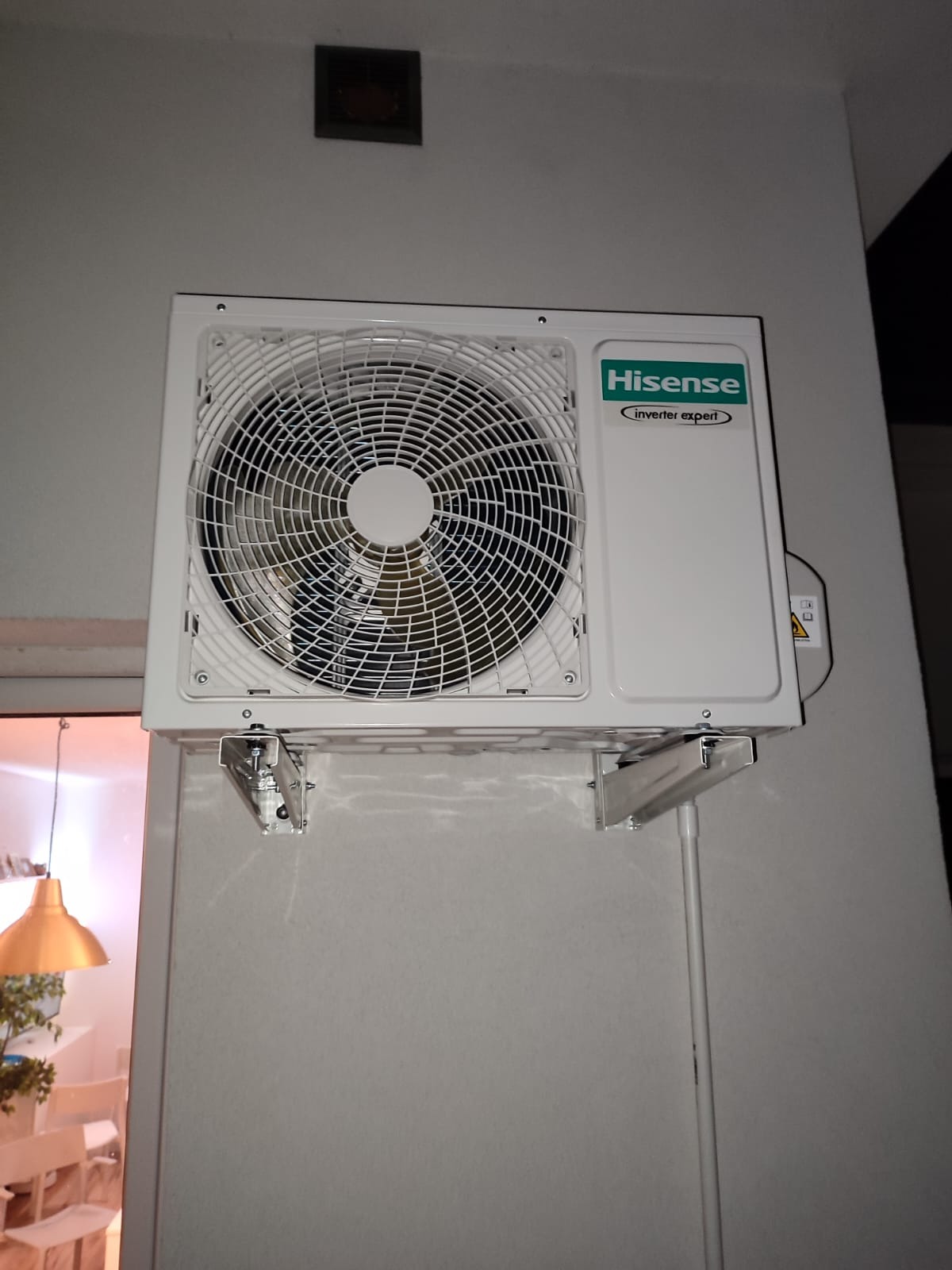 Zewnętrzna jednostka klimatyzacji Hisense zamontowana na szarej ścianie, widoczne metalowe wsporniki i biała rura instalacyjna, w tle fragment pomieszczenia z lampą.