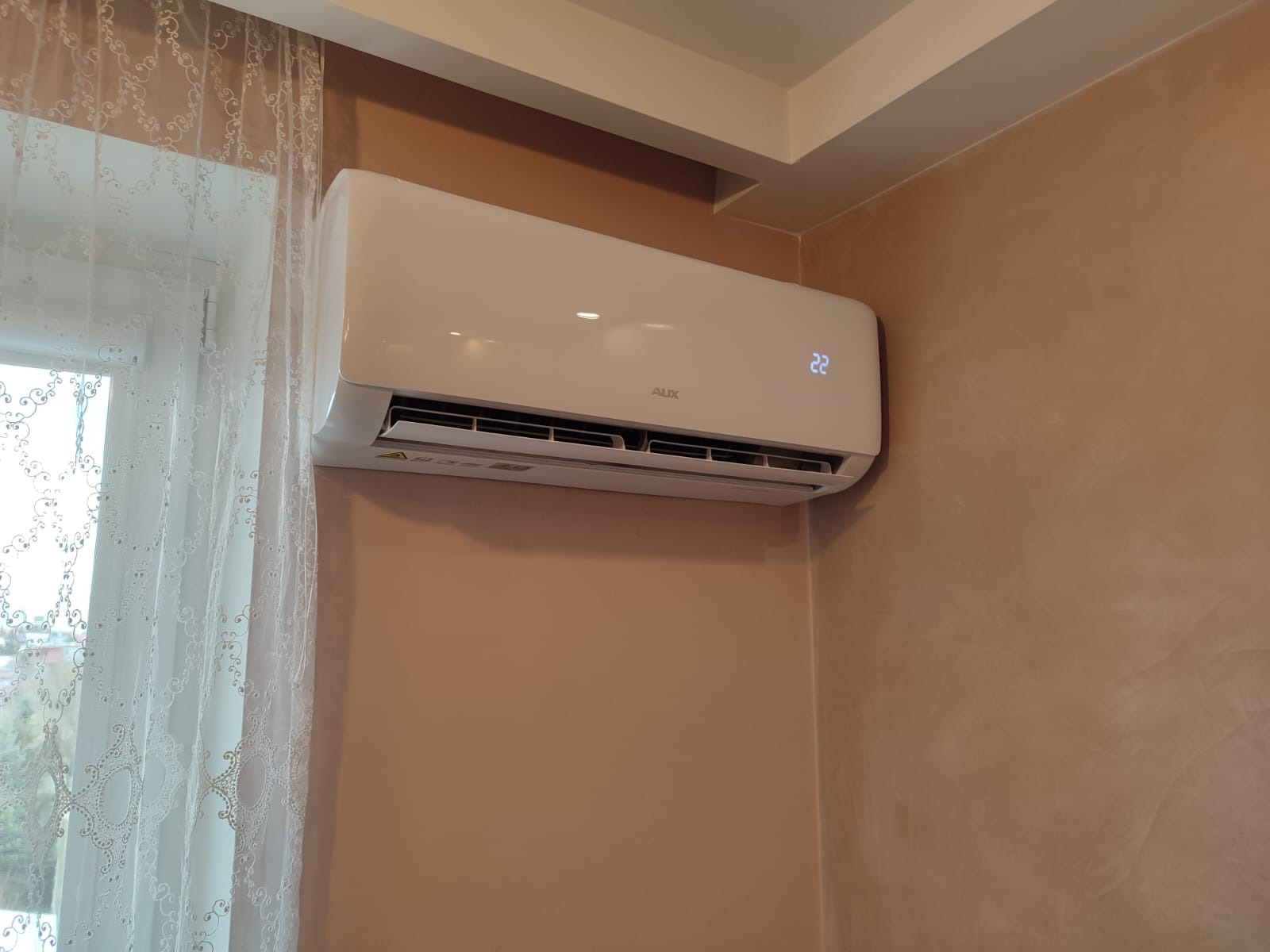 Biały klimatyzator marki AUX zamontowany na beżowej ścianie, obok okna z firanką. Na wyświetlaczu klimatyzatora widoczna temperatura 22 stopnie.
