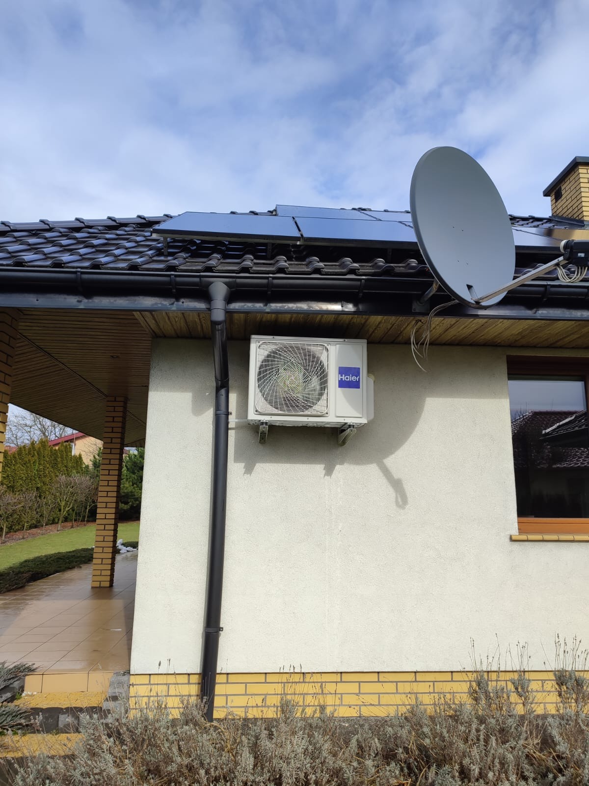 Biała jednostka zewnętrzna klimatyzacji Haier zamontowana na ścianie domu z brązowymi panelami fotowoltaicznymi na dachu i anteną satelitarną.