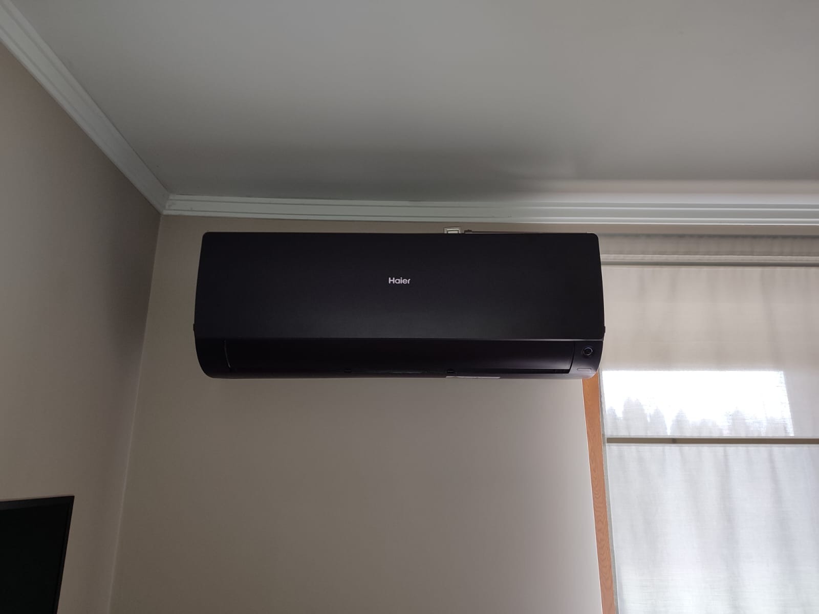 Czarny klimatyzator Haier zamontowany na ścianie w pomieszczeniu z kremowymi ścianami i białym sufitem z dekoracyjną listwą. Obok okno z delikatnymi, jasnymi roletami.