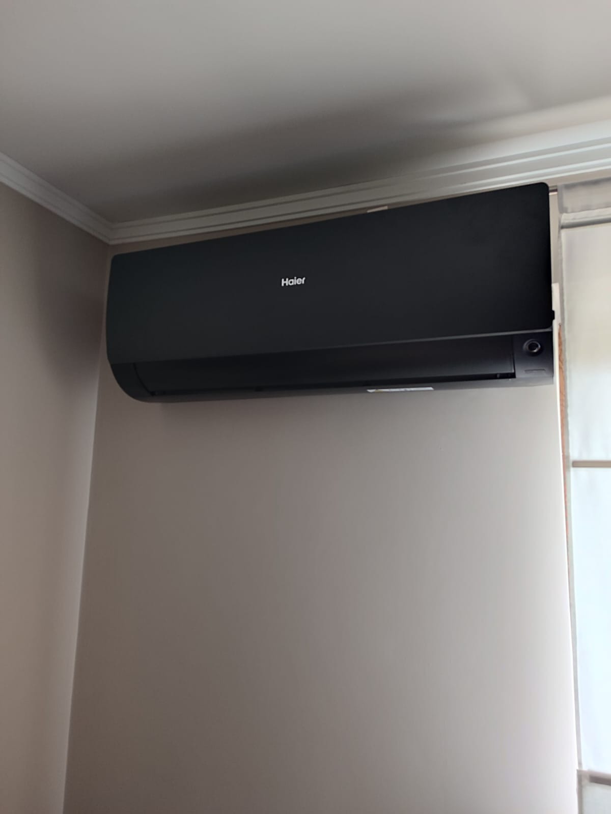 Czarny klimatyzator Haier zamontowany na beżowej ścianie w pomieszczeniu z widoczną białą listwą sufitową i zasłoną w tle.