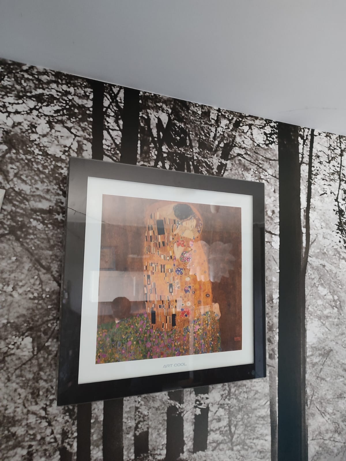 Oprawiony w czarną ramę obraz 'Pocałunek' Gustava Klimta wisi na ścianie pokrytej tapetą z motywem czarno-białego lasu, widoczny napis ART COOL na dole ramki.