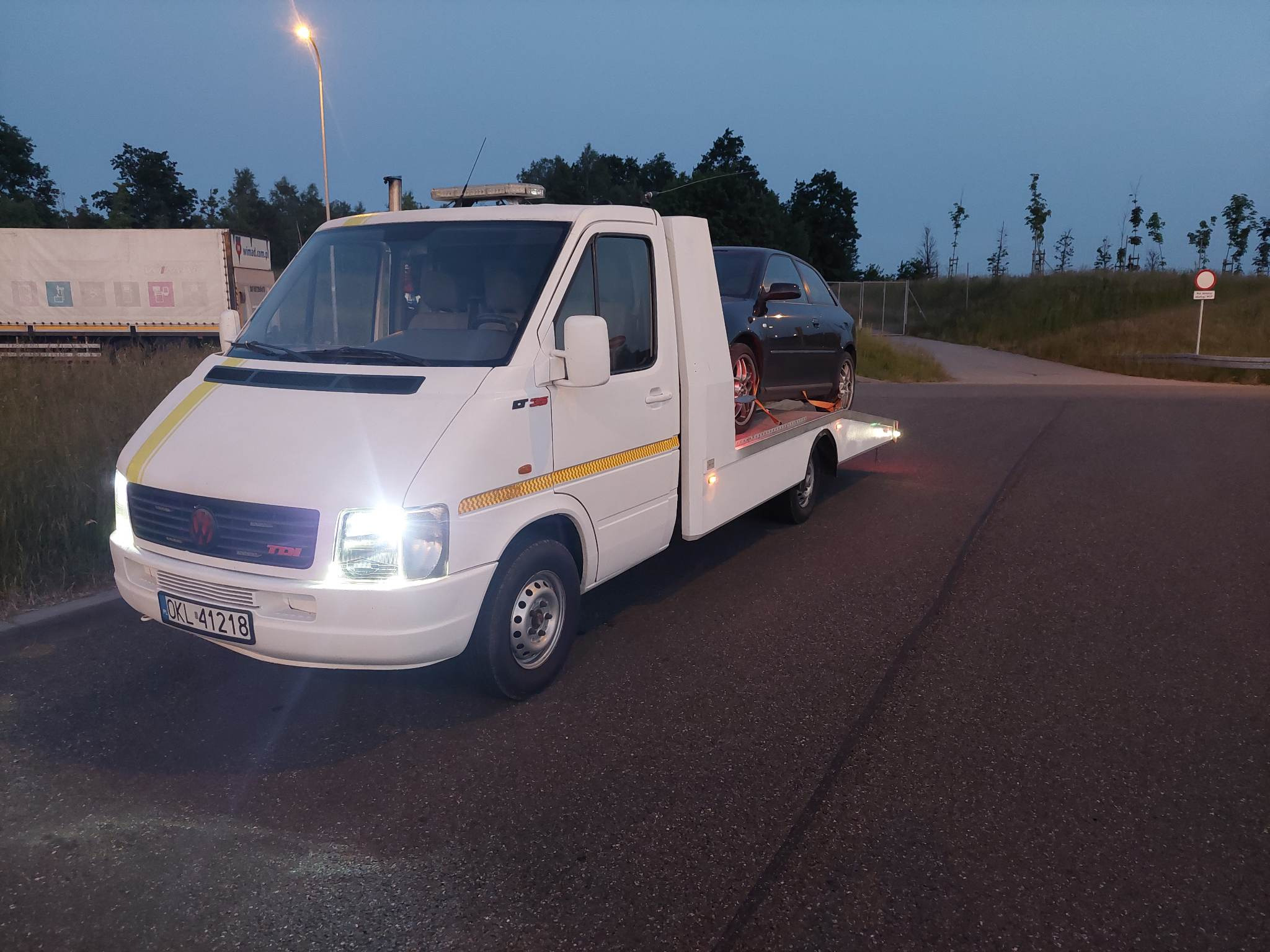 Biała laweta z zapalonymi światłami przewozi ciemny samochód osobowy na tle drogi i drzew przy zmierzchu.