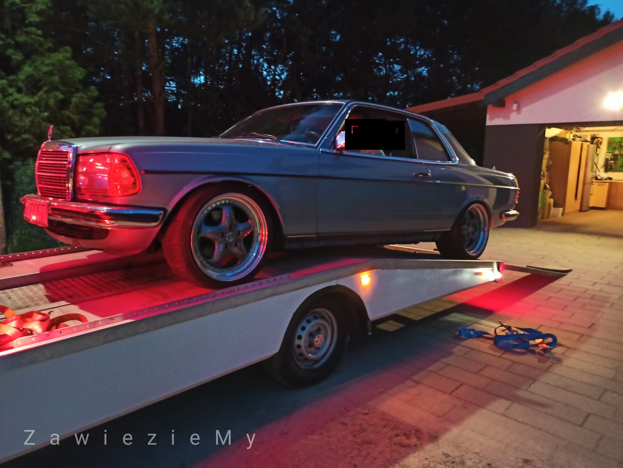 Niebieski Mercedes-Benz W123 na białej lawecie oświetlonej czerwonym światłem, widoczne pasy mocujące i garaż w tle.