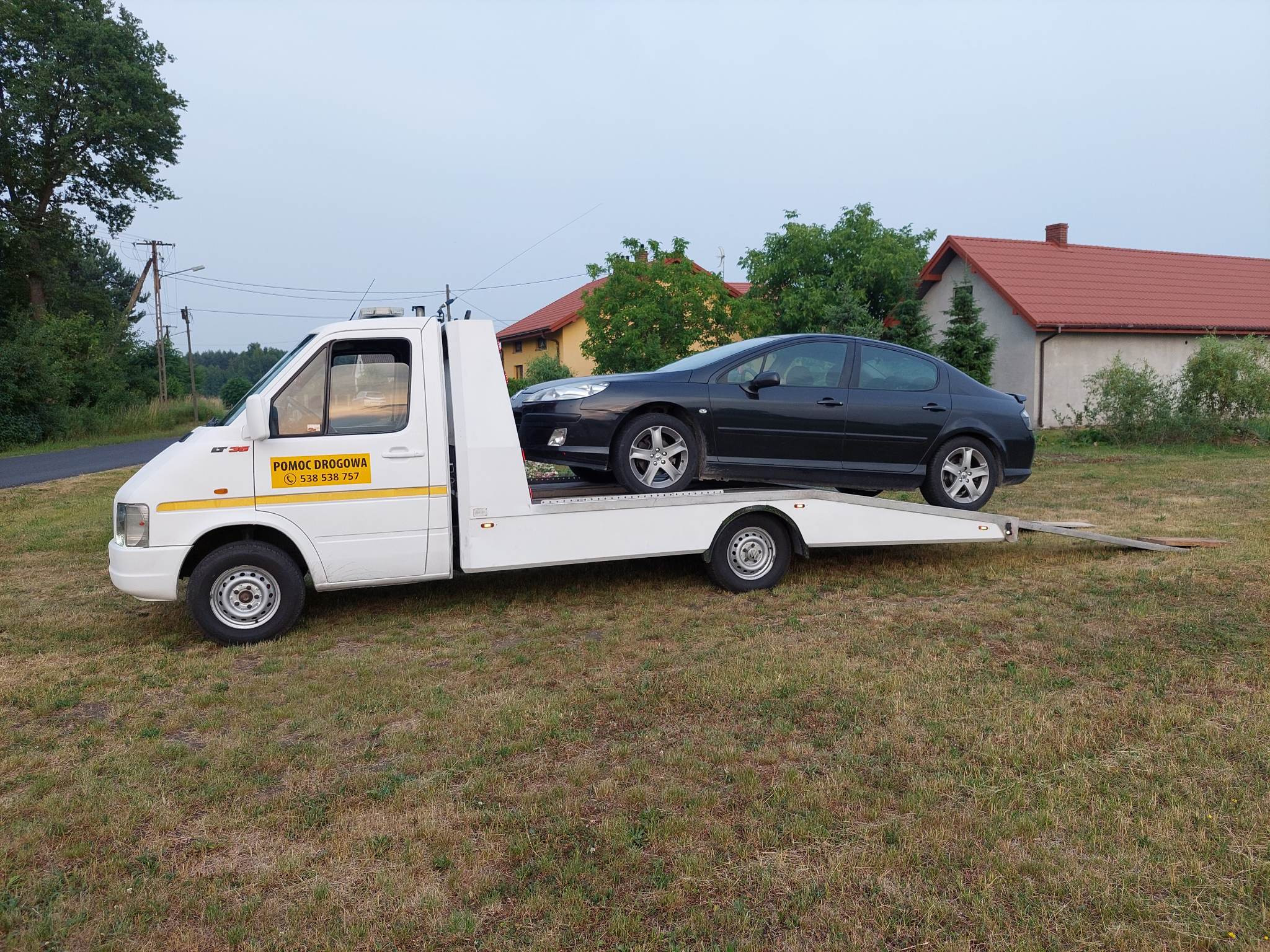 Czarny samochód osobowy Peugeot na białej lawecie pomocy drogowej z żółtym paskiem i napisem 'Pomoc Drogowa' na tle wiejskiego krajobrazu z domami.