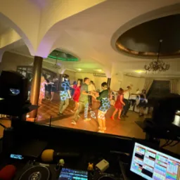 Parkiet taneczny z bawiącymi się ludźmi, widziany zza konsolety DJ'a. Oświetlenie punktowe tworzy wzory na ubraniach tancerzy. W tle kolumny i żyrandole.