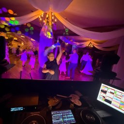 VENOM WORKS WŁADYSŁAW CHWASTEK - DJ grający na weselu, widok zza konsoli na parkiet z tańczącymi gośćmi. Sufit zdobią białe materiały i kolorowe światła. Na pierwszym planie sprzęt DJ-ski Numark.