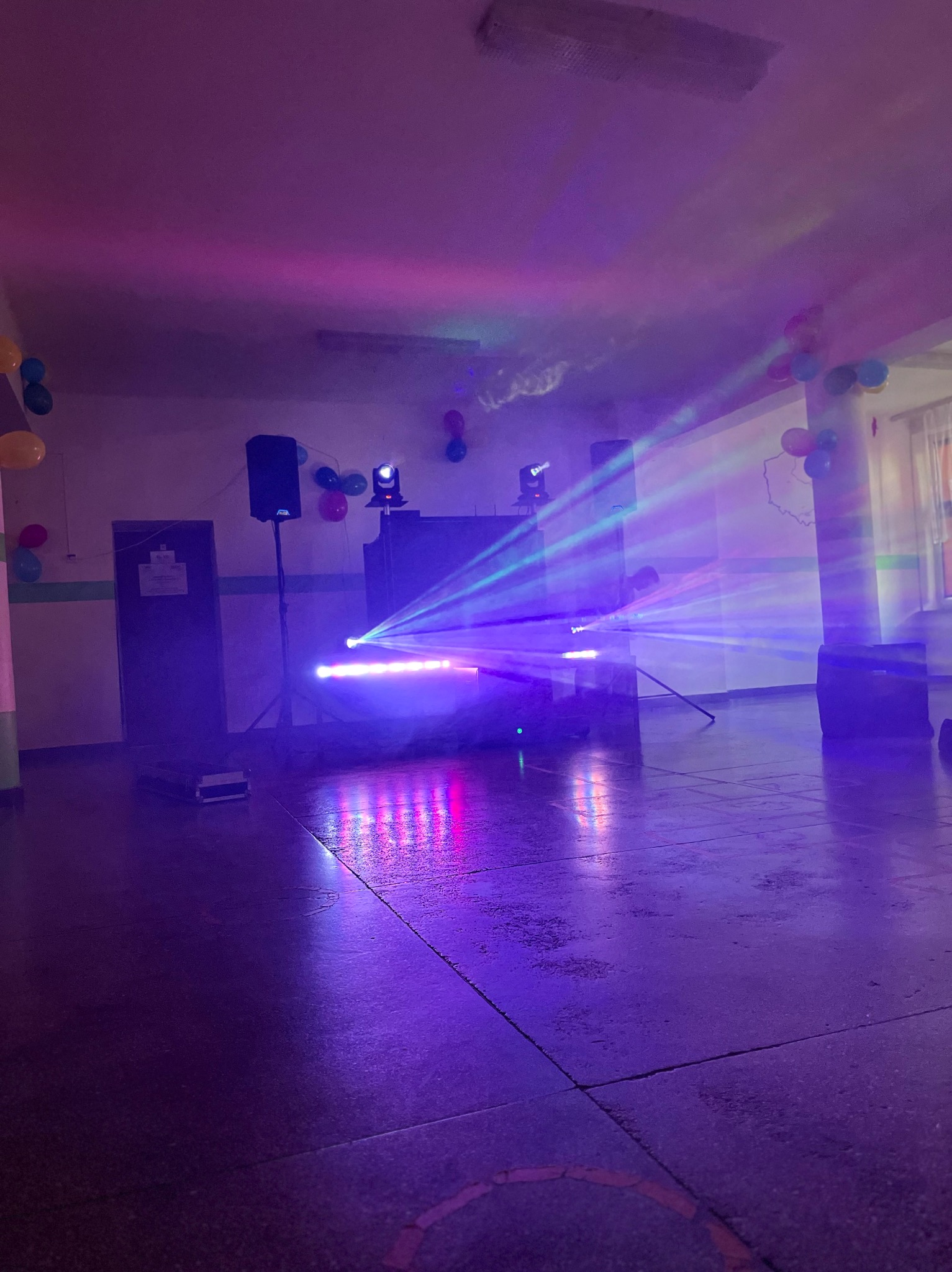Sala z nagłośnieniem, oświetleniem i balonami, przygotowana do imprezy. Widoczne reflektory, głośniki i konsoleta DJ-ska. Fioletowe światło wypełnia przestrzeń.