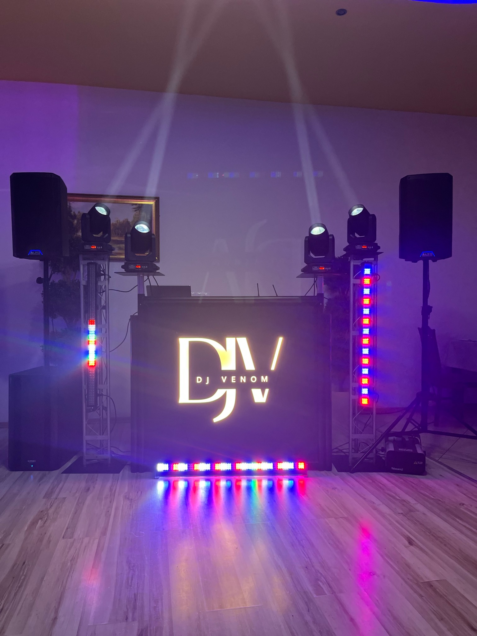 Stanowisko DJ'a z logo 'DJ Venom' podświetlone kolorowymi światłami, z kolumnami głośnikowymi i reflektorami na metalowych konstrukcjach, ustawione na drewnianej podłodze.