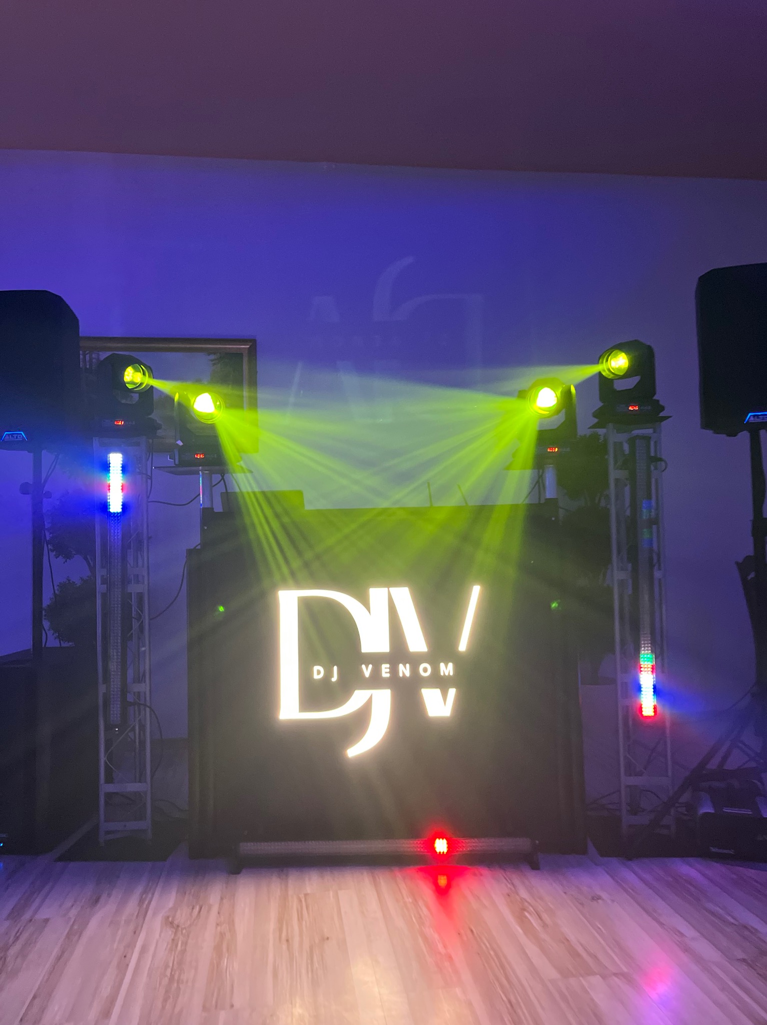 Scena DJ-a z oświetleniem: reflektory z zielonymi promieniami, panel z logo 'DJ Venom' podświetlony na biało, kolumny głośnikowe po bokach, parkiet podświetlony na różowo-fioletowo.