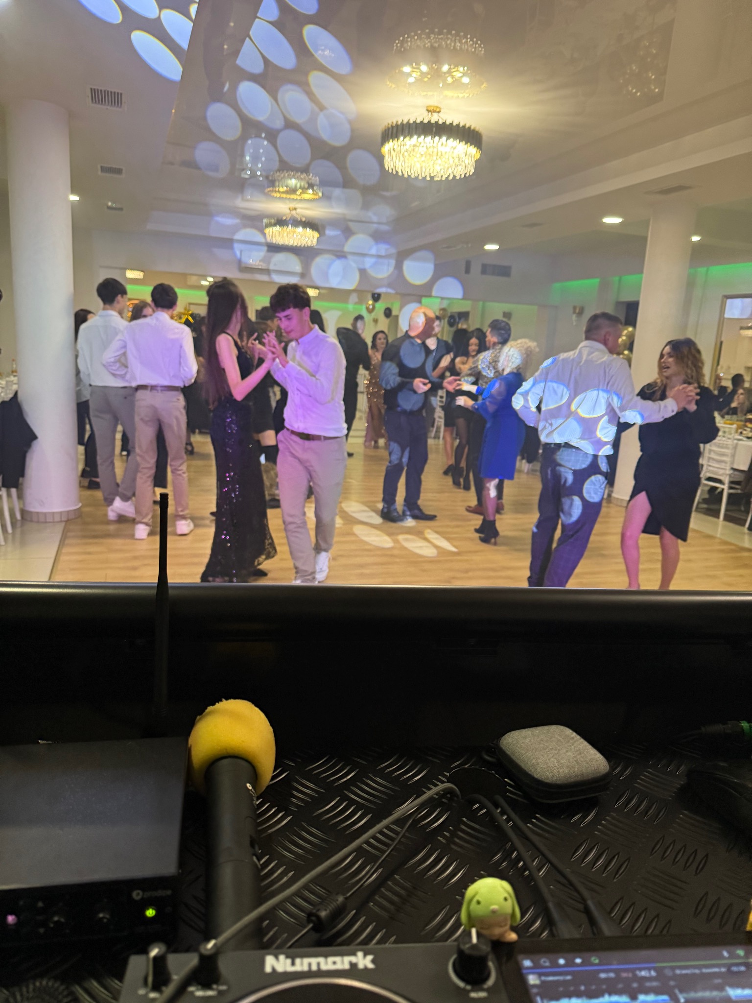 Parkiet taneczny z bawiącymi się gośćmi, widziany z perspektywy stanowiska DJ-a z mikrofonem i sprzętem marki Numark. Oświetlenie punktowe na suficie i ścianach.