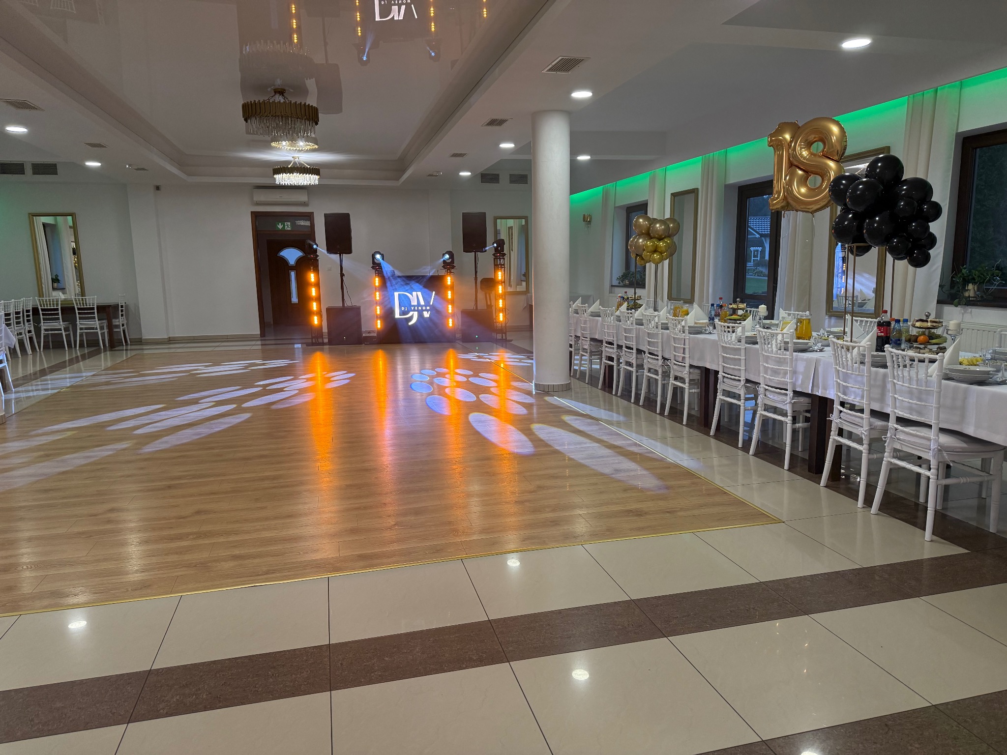 Sala balowa z nagłośnieniem, oświetleniem i dekoracją na 18 urodziny. Parkiet oświetlony reflektorami, stoły z białymi obrusami i krzesłami.
