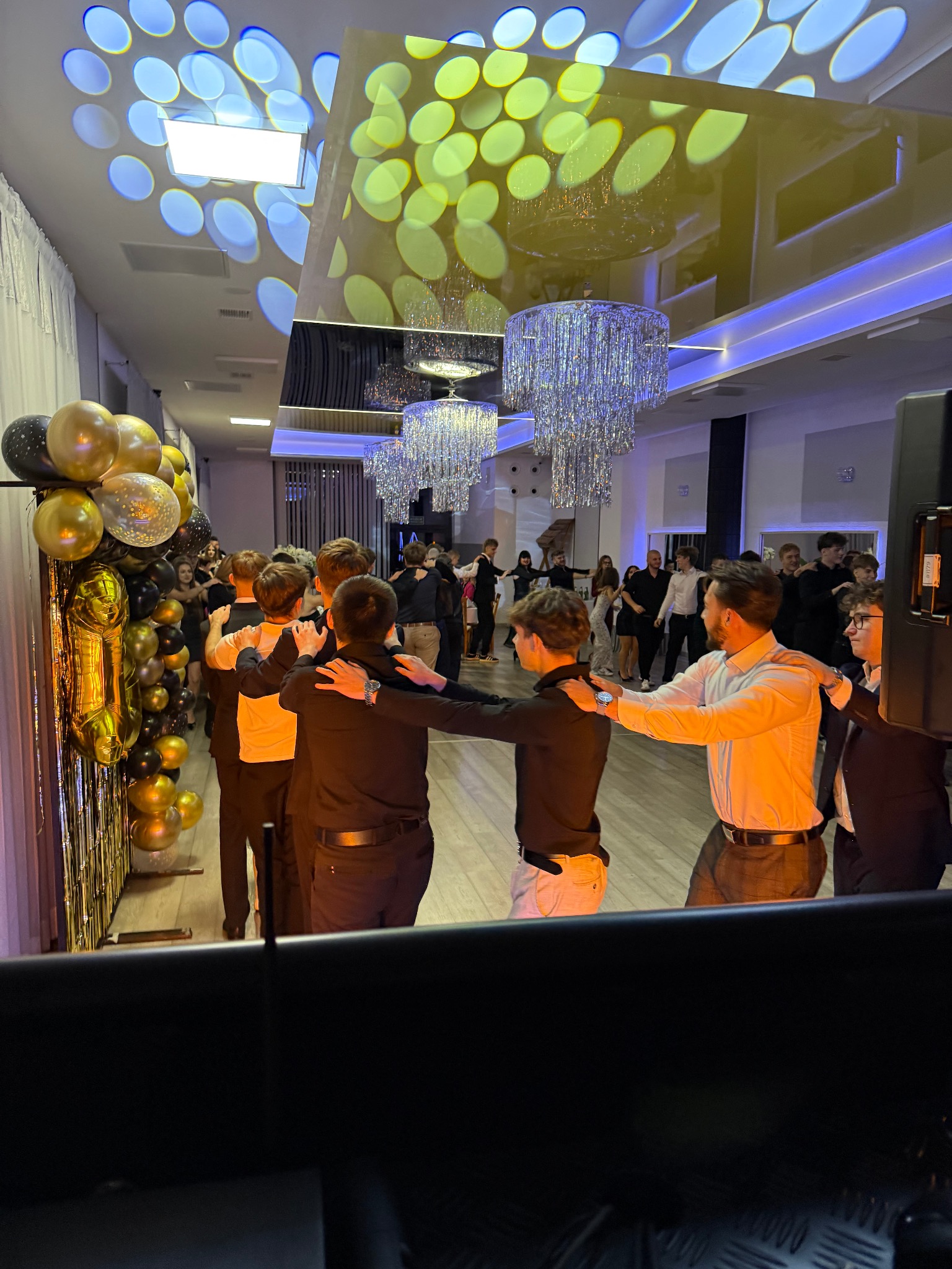 Uczestnicy studniówki w trakcie poloneza, dekoracje z balonów w odcieniach złota i czerni, oświetlenie parkietu z efektem bokeh.