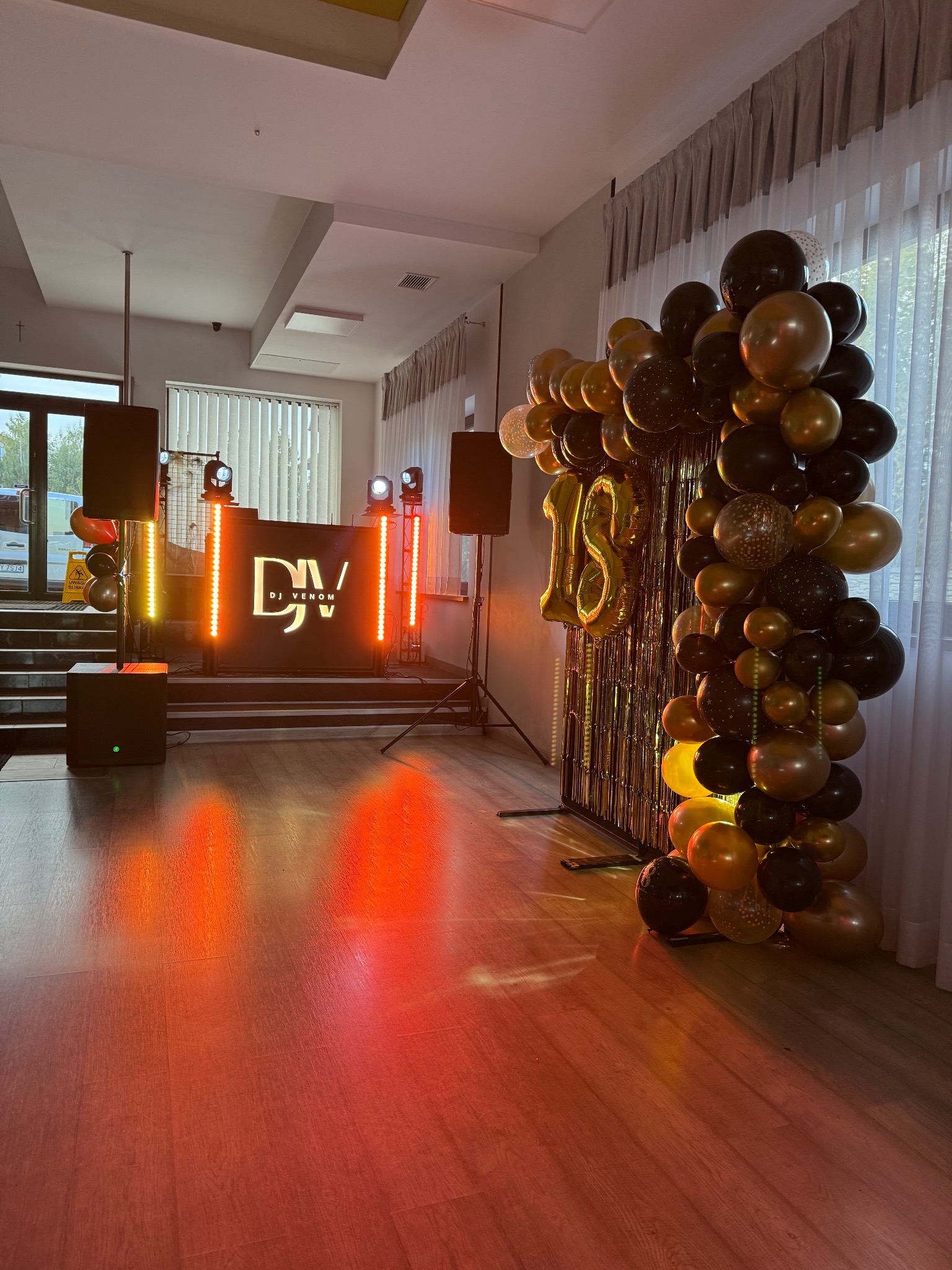 Dekoracja sali na 18 urodziny: czarno-złote balony, kurtyna, podświetlane stanowisko DJ-a z logo 'DJ Venom', parkiet oświetlony na czerwono.