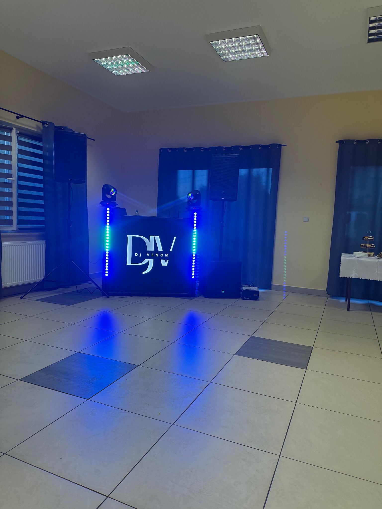 Ustawiony sprzęt DJ'ski z podświetleniem LED w sali z oknami i zasłonami. Widoczne głośniki, mikser, oświetlenie i logo 'DJ Venom'.  Podłoga wyłożona płytkami.