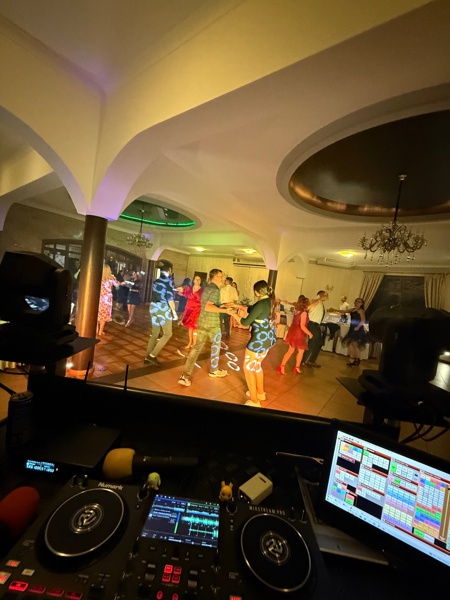 Parkiet taneczny z bawiącymi się ludźmi, widziany zza konsolety DJ'a. Oświetlenie punktowe tworzy wzory na ubraniach tancerzy. W tle kolumny i żyrandole.