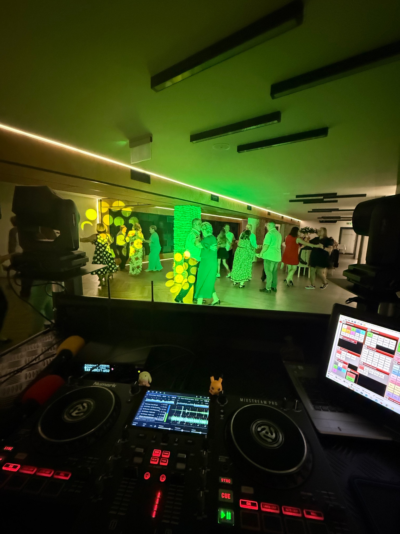 DJ na weselu: parkiet pełen tańczących gości oświetlony zielonymi światłami, widok zza konsoli DJ-skiej Numark Mixstream Pro z laptopem.