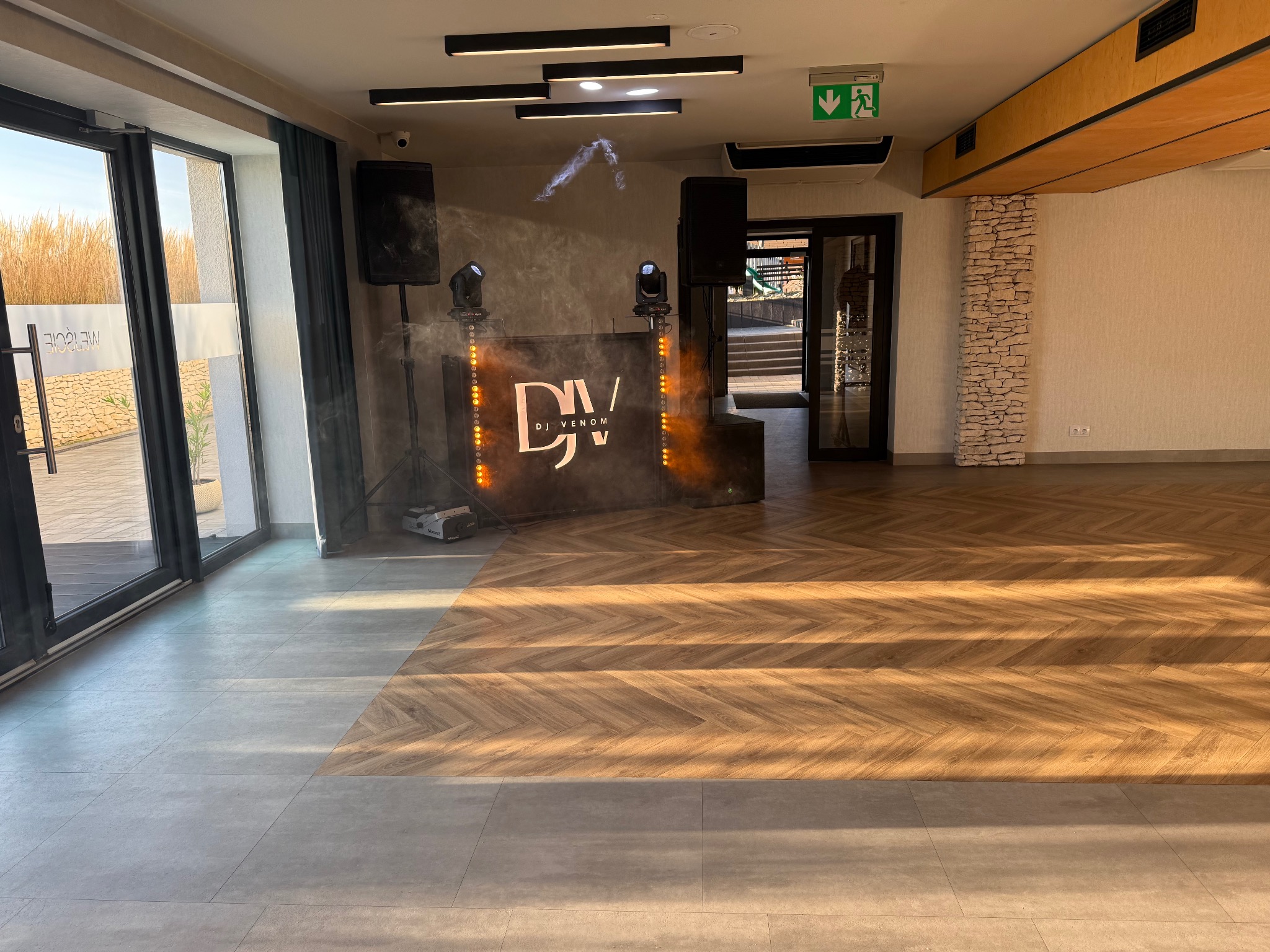 Sala eventowa z parkietem w jodełkę, oświetleniem scenicznym i stanowiskiem DJ'a z logo 'DJ Venom'. Widoczne głośniki i światła. Przygotowania do imprezy.