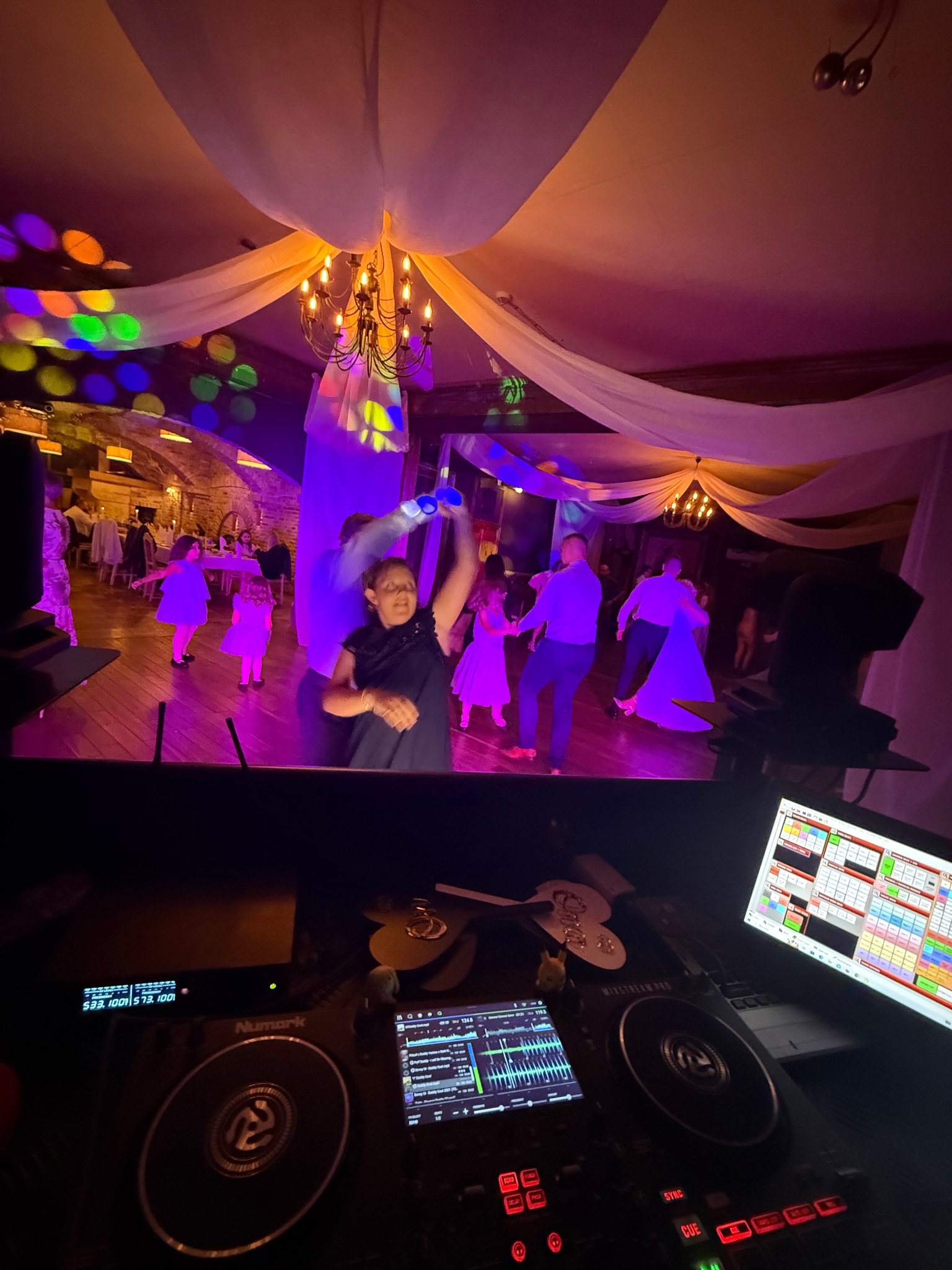 DJ grający na weselu, widok zza konsoli na parkiet z tańczącymi gośćmi. Sufit zdobią białe materiały i kolorowe światła. Na pierwszym planie sprzęt DJ-ski Numark.