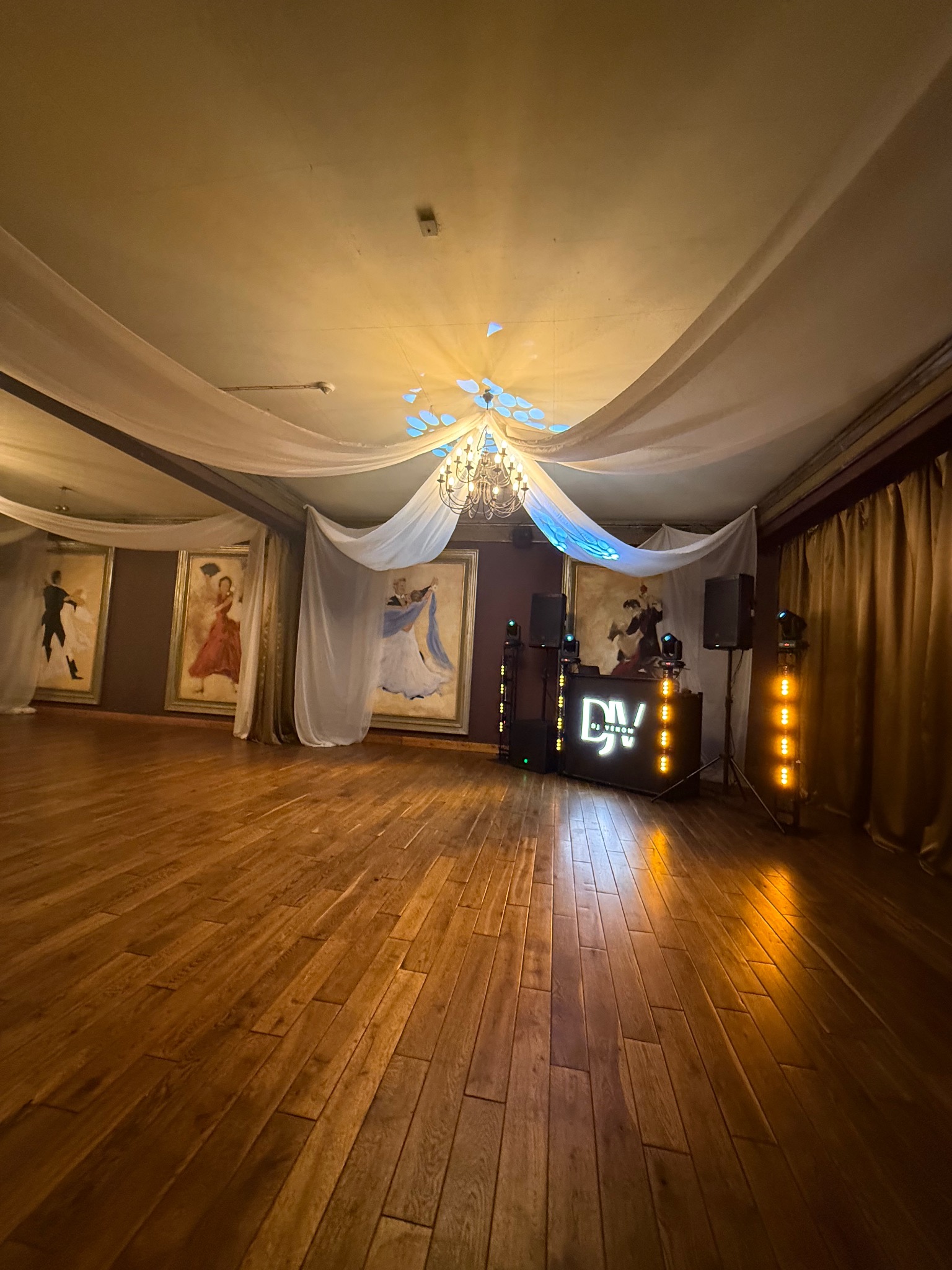 Sala balowa z parkietem, dekoracją z białego materiału, oświetleniem DJ-a i obrazami tancerzy. Gotowa na imprezę taneczną.