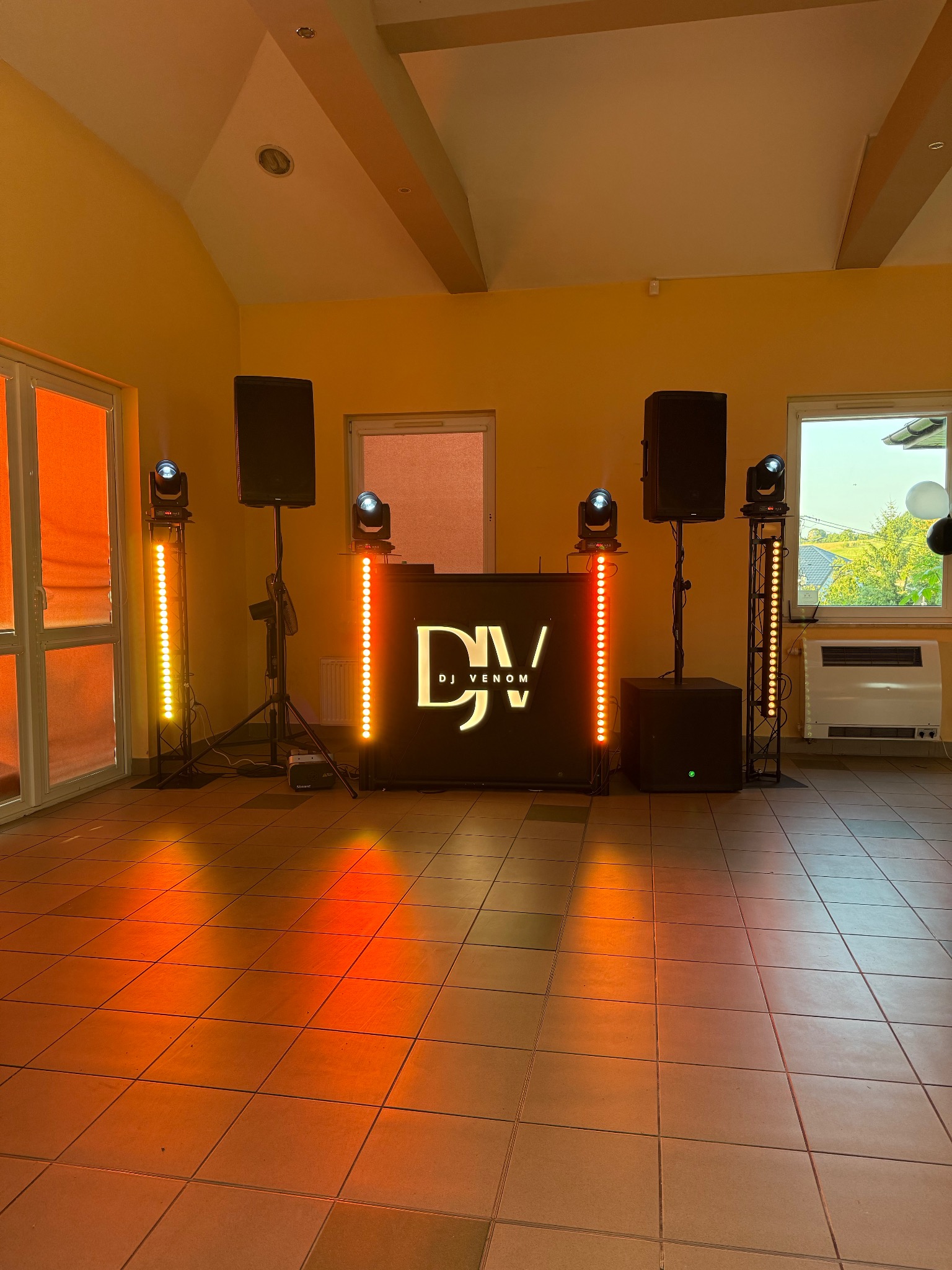 Kompletny zestaw nagłośnieniowy DJ Venom z oświetleniem LED w pomarańczowym pomieszczeniu z oknami. Sprzęt gotowy do imprezy, widoczne kolumny, subwoofer i oświetlenie.