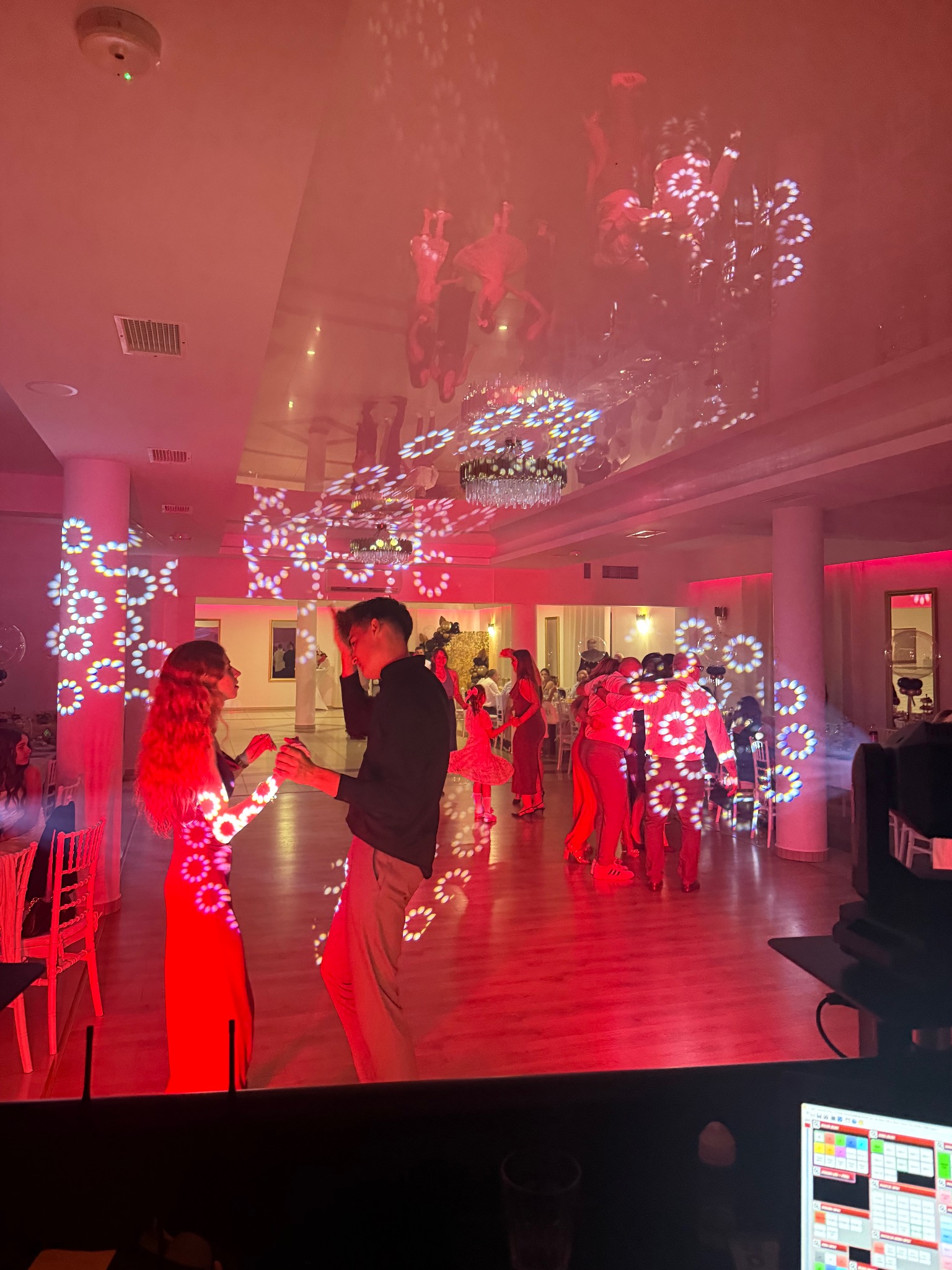 Para tańczy na parkiecie oświetlonym czerwonym światłem i wzorami reflektorów podczas imprezy. W tle widoczni inni ludzie i elementy wystroju sali.