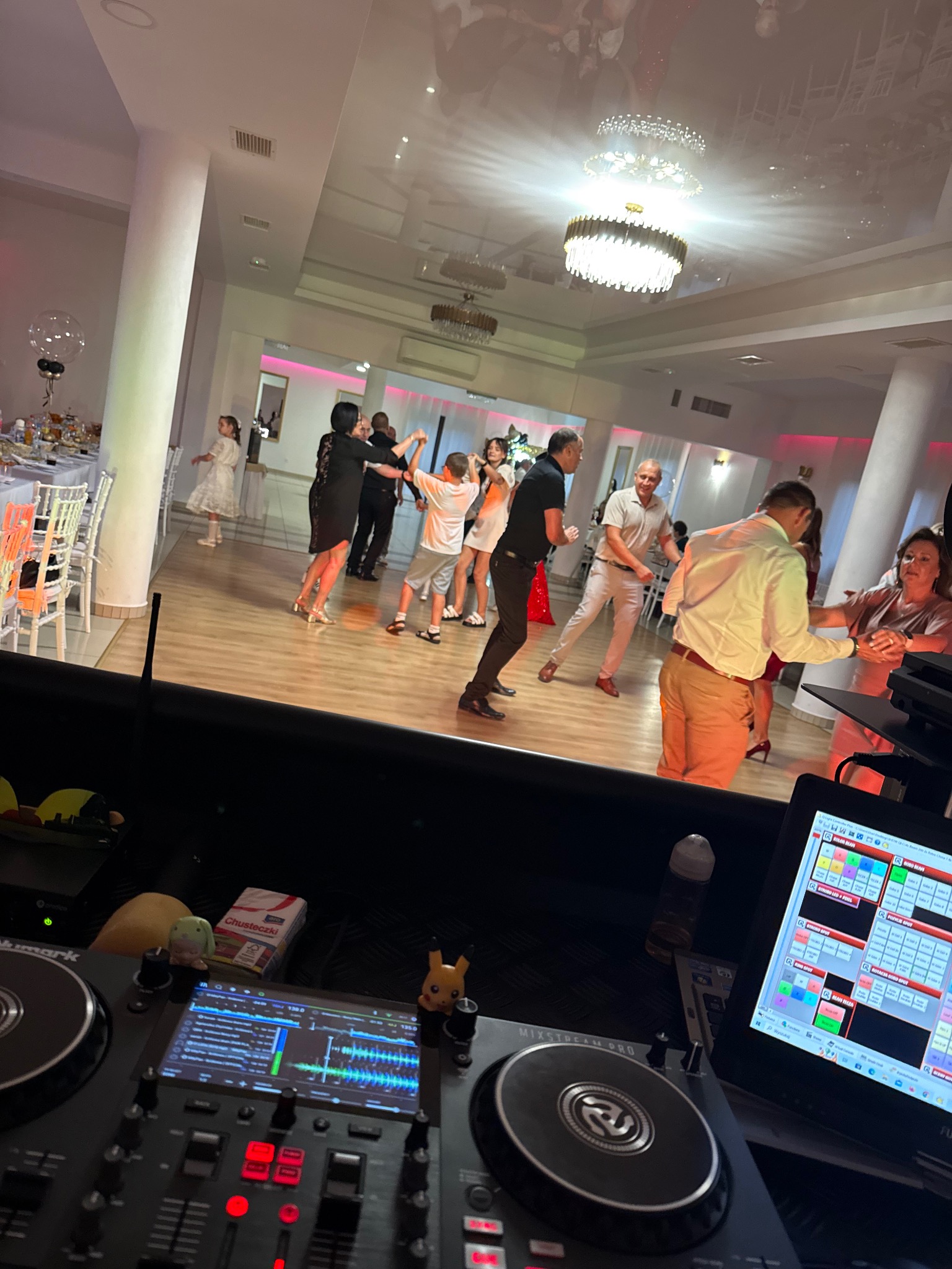 Parkiet taneczny z bawiącymi się gośćmi widziany z perspektywy DJ'a, z widocznym sprzętem DJ'skim i laptopem. W tle dekoracje sali weselnej.